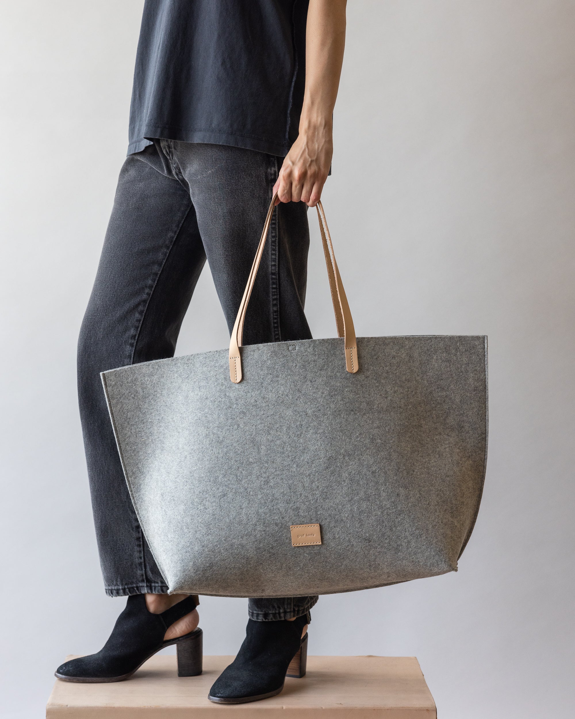 A person in a black shirt, dark jeans, and black heeled shoes holds a large graf lantz gray felt tote bag with tan leather handles. They stand on a wooden platform against a plain background, showcasing effortless style ideal for casual beach days.