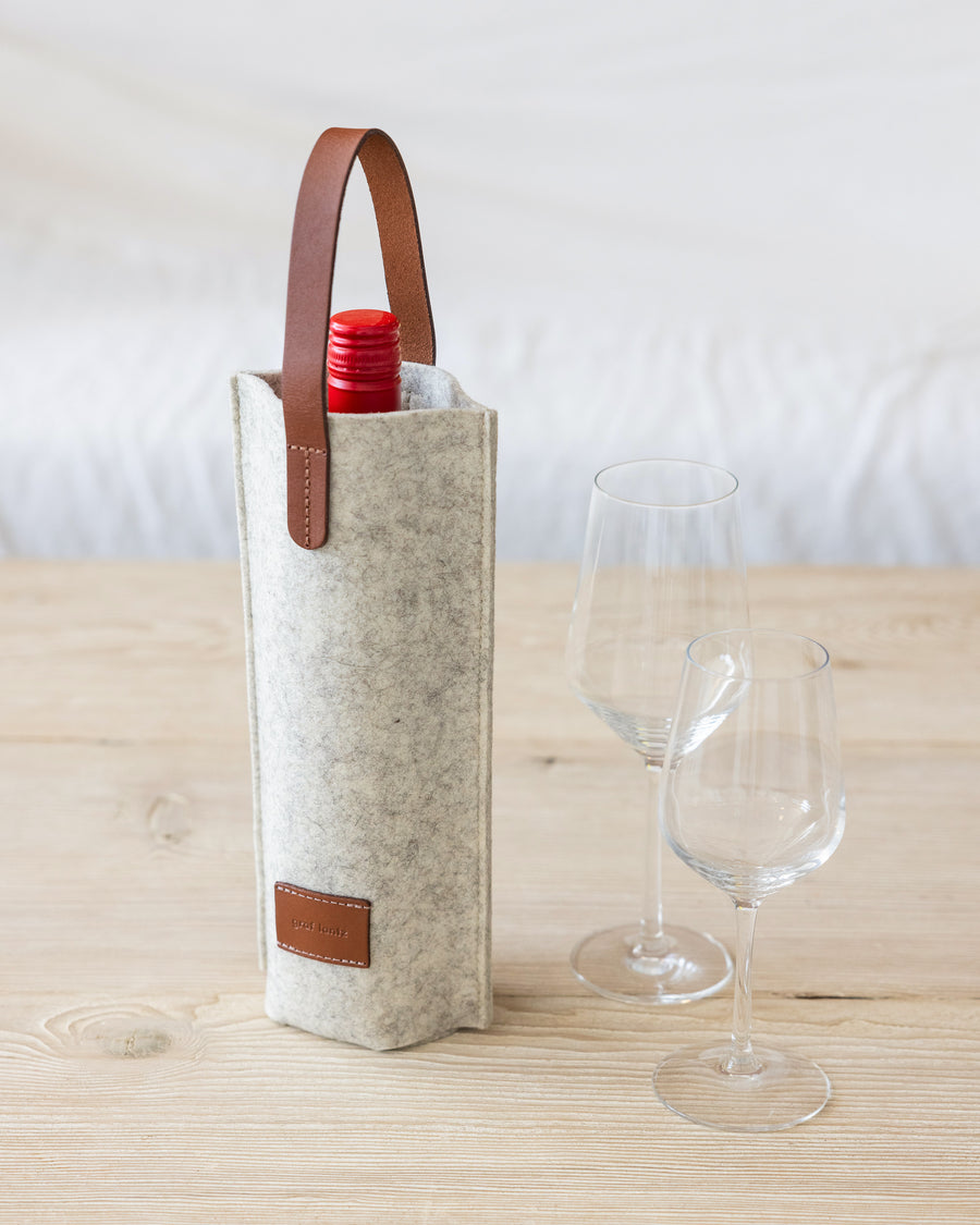 A wine bottle in a white Merino Wool Felt Single Wine Carrier by Graf Lantz with a brown leather handle on a wooden tabletop 