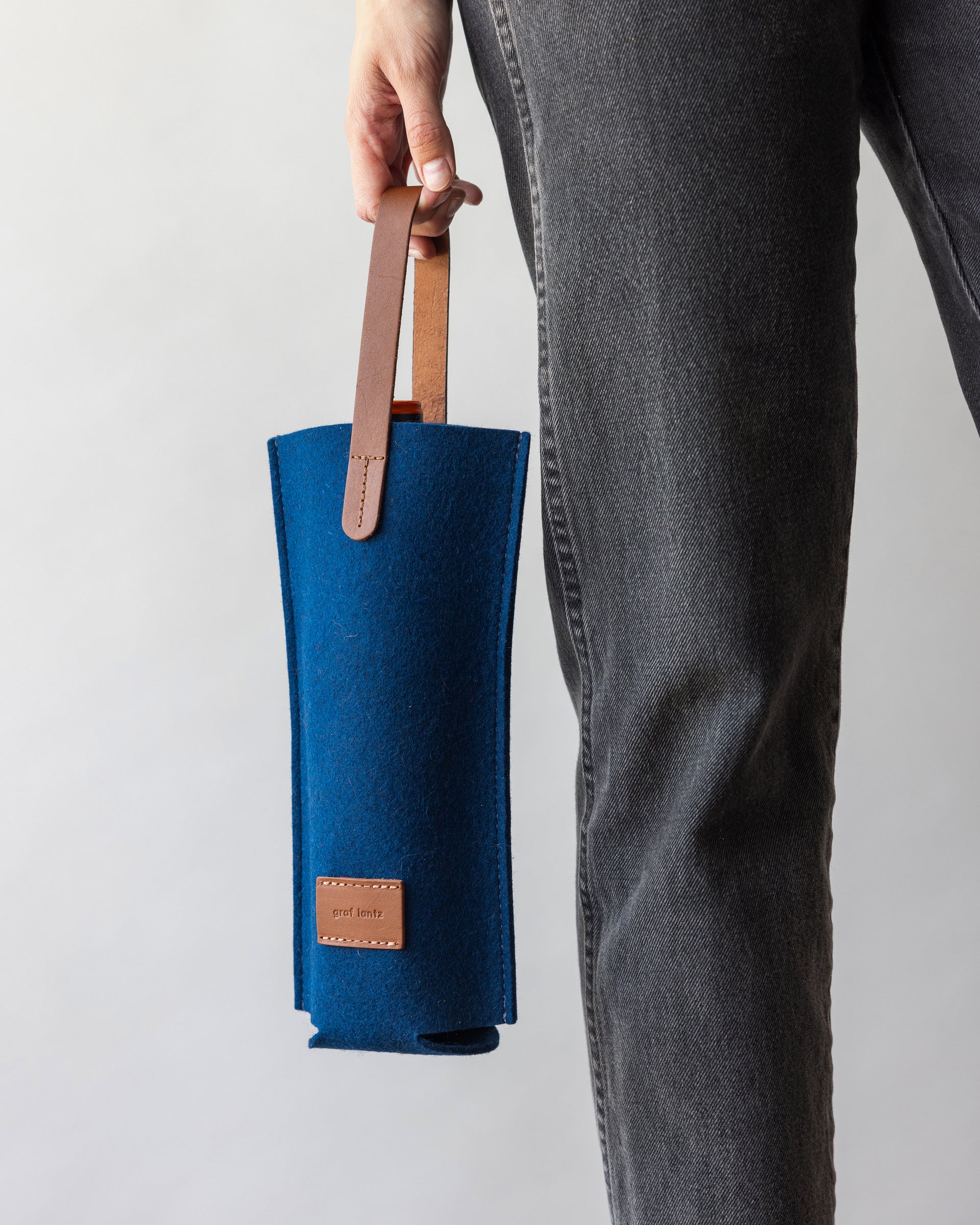 A woman holding a Merino Wool Felt Single Wine Carrier in blue with a brown leather handle, front view