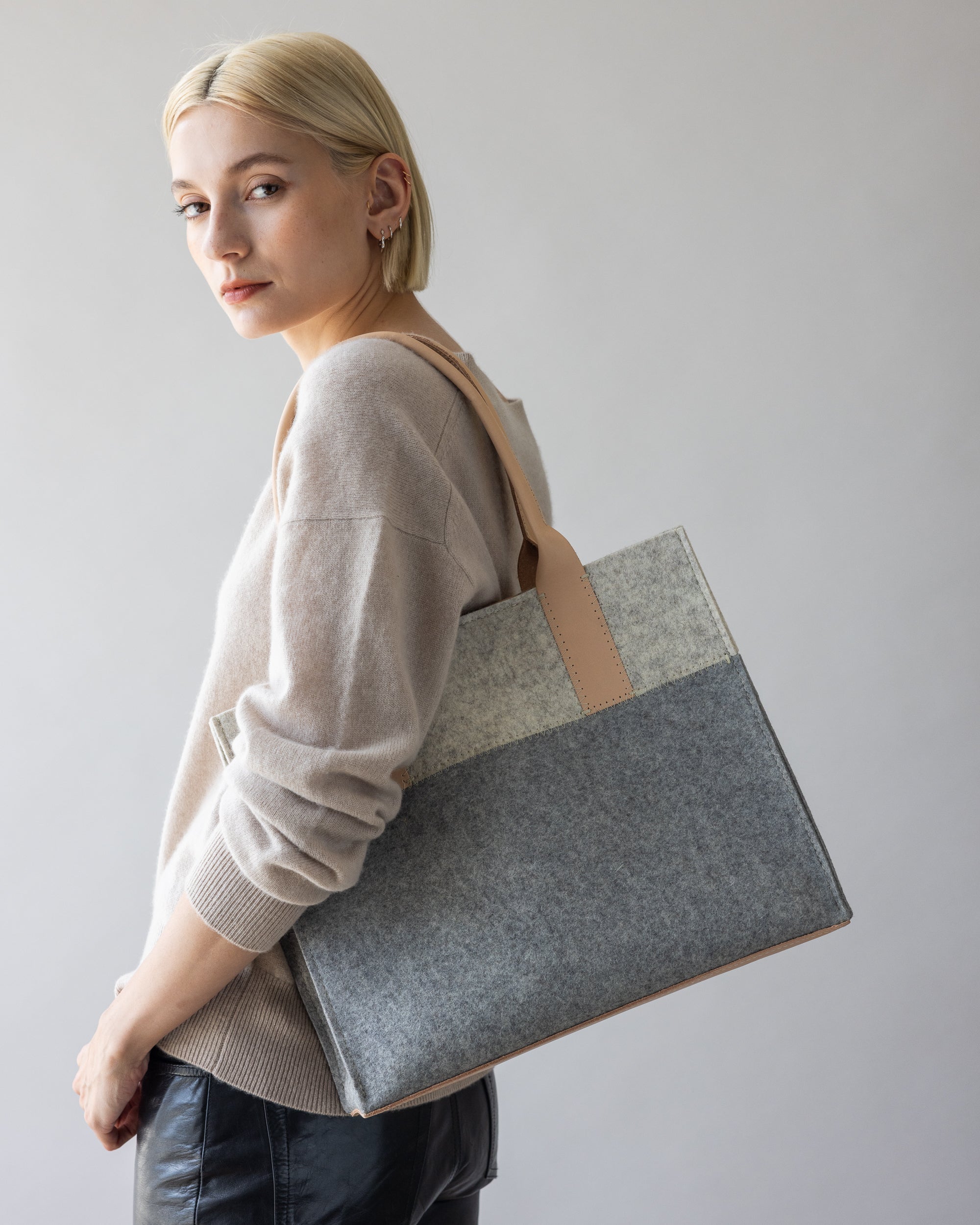 A person with short blonde hair wears a beige sweater and black pants, looking sideways. They carry a stylish graf lantz Jaunt Midi Tote in grey and beige over their shoulder. Soft lighting enhances the serene scene.
