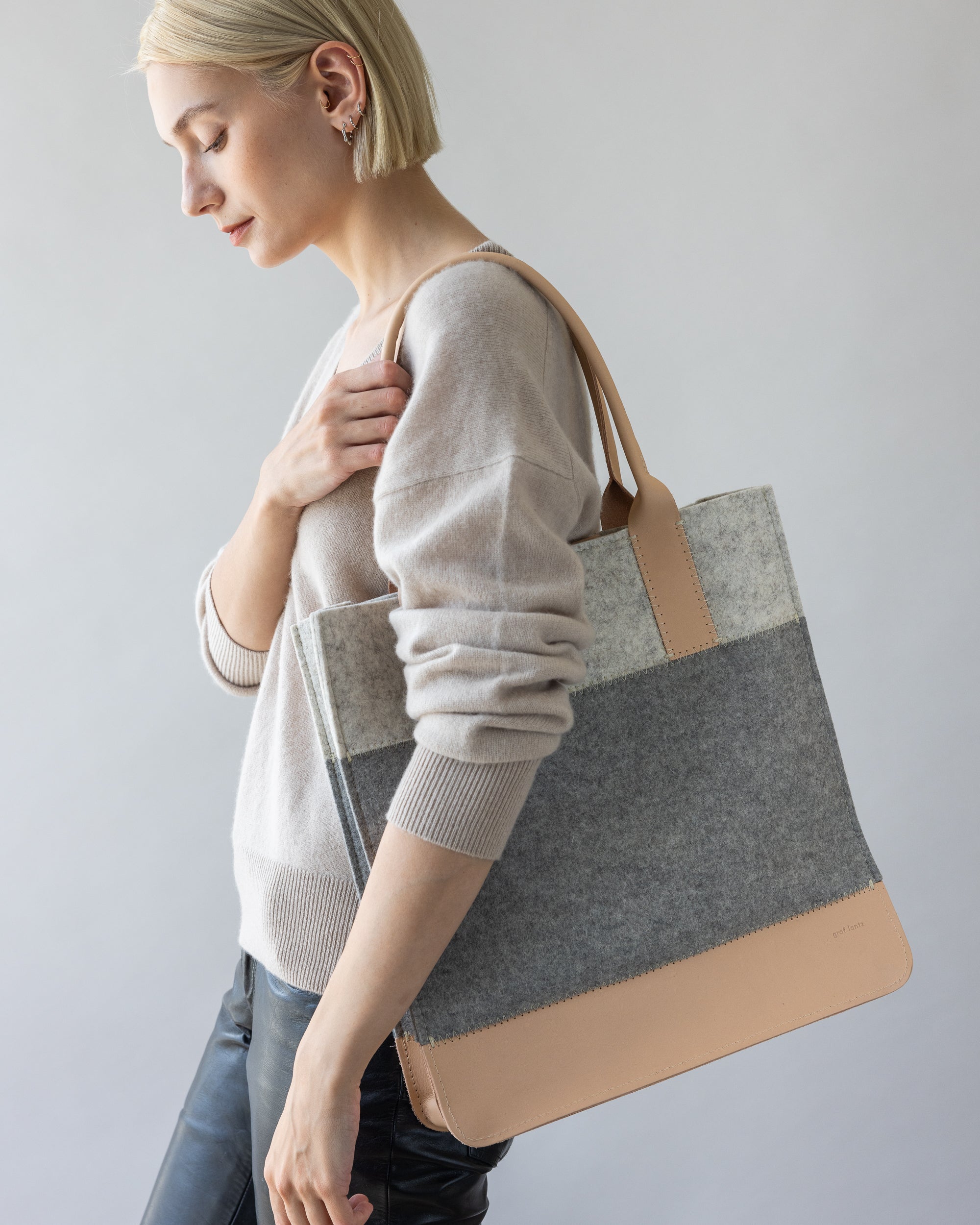 A person with short blond hair stands against a neutral backdrop, holding a large gray and tan tote by graf lantz. Theyre dressed in a light beige Merino wool sweater and black pants, exuding thoughtful sustainable elegance.