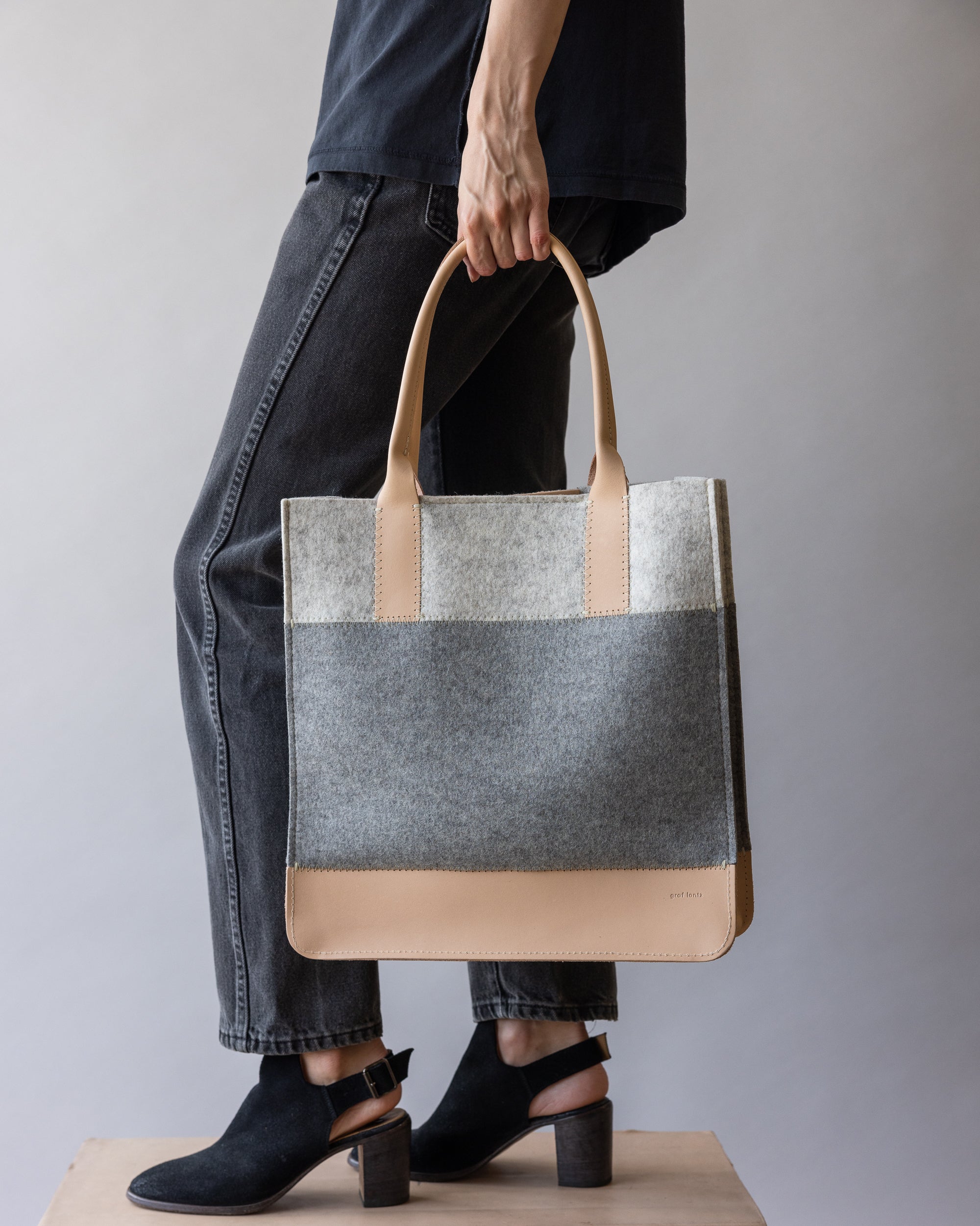 A model carries a graf lantz sustainable tote bag with gray and beige panels, paired with black jeans, a black shirt, and black heeled ankle boots against a neutral gray backdrop.