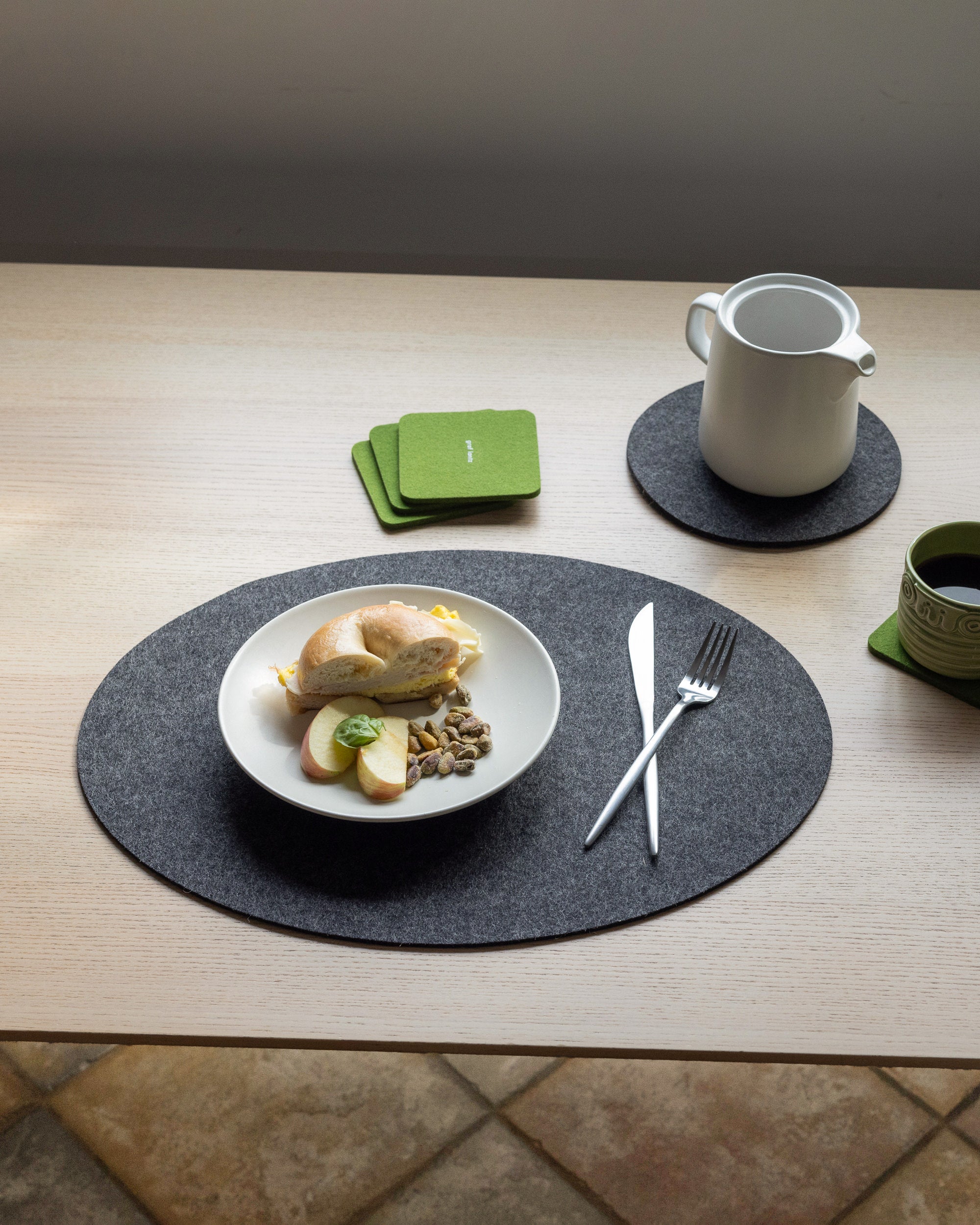 A breakfast setting on a light wooden table showcases graf lantz Oeko-Tex® Certified Merino wool placemat with bread, cheese, apple slices, and nuts. Silverware is nearby, complemented by a white pitcher, green coasters from graf lantz, and a matching green cup.