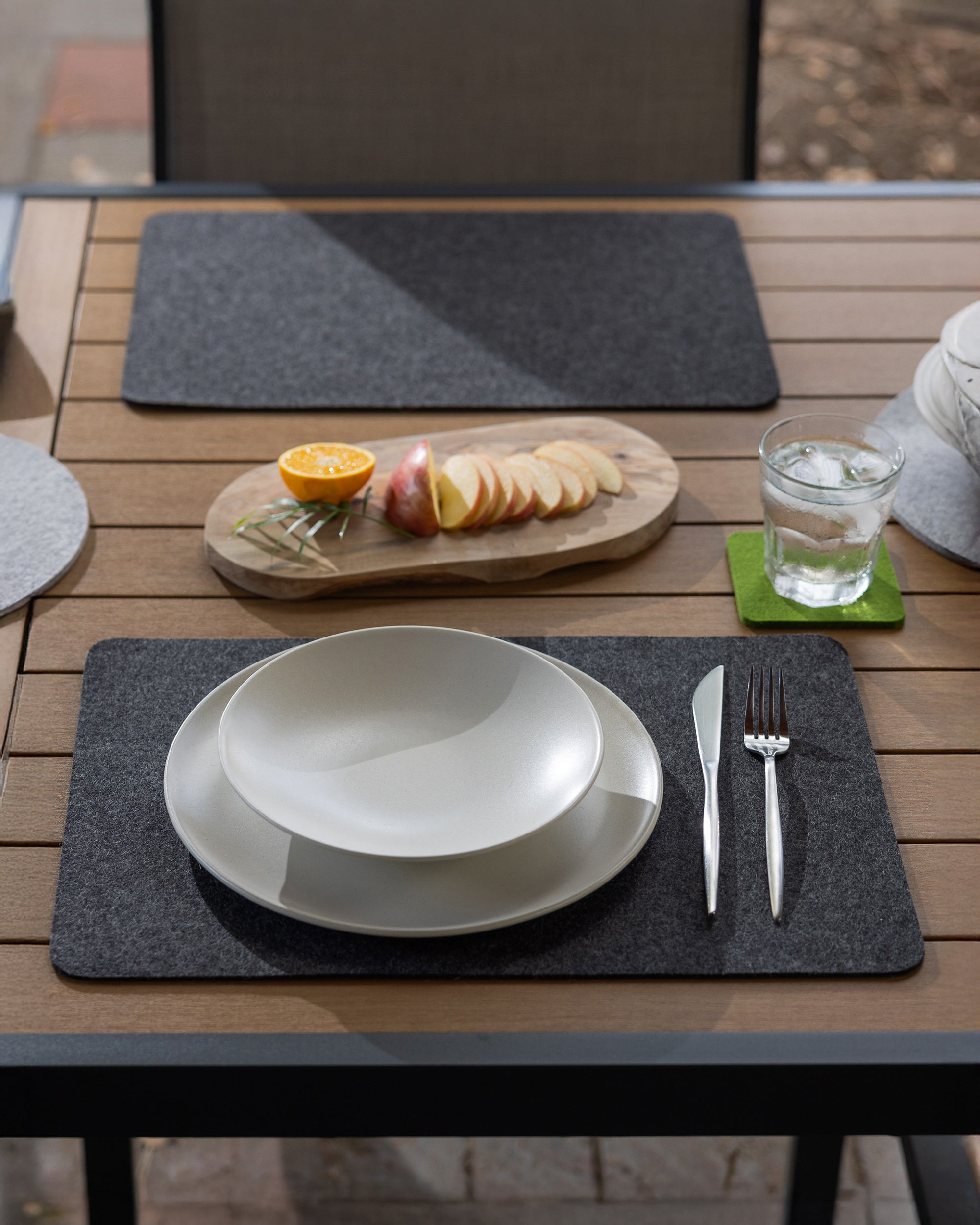 An outdoor table setting featuring a graf lantz Merino Wool Placemat, white bowl and plate, silverware, and a glass of water. Sliced apple and half an orange rest on a wooden board, with the moisture-wicking placemat elegantly set on the patio.