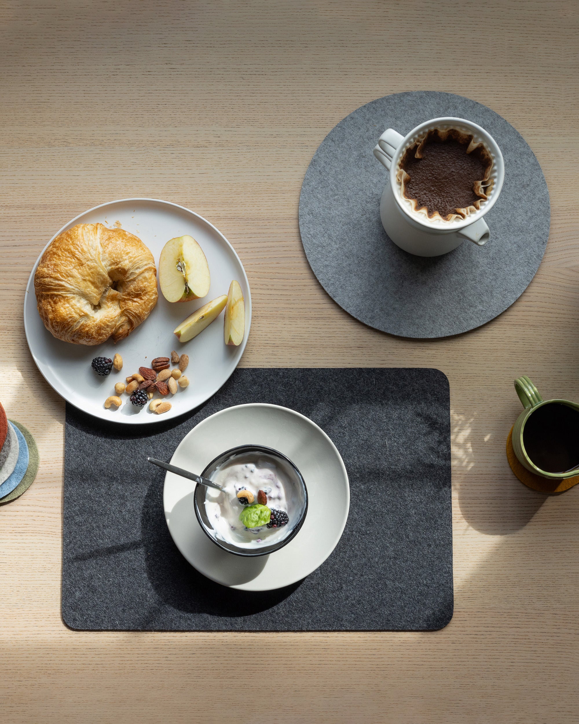 A breakfast setting on a wooden table includes a white plate with a croissant, apple slices, nuts, and berries. Its complemented by black coffee and kiwi-blackberry yogurt, all on graf lantz Merino Wool Placemats, both round and square in shape.