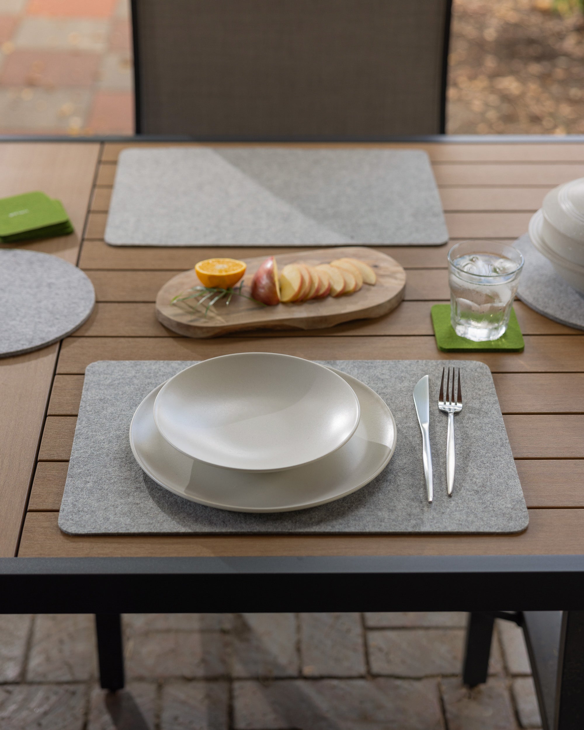 The outdoor dining table set includes graf lantz Merino Wool placemats, a white stacked bowl and plate, a knife and fork, and a water glass. A wooden board with sliced apples and an orange half sits on top. Stone paving is visible underneath.