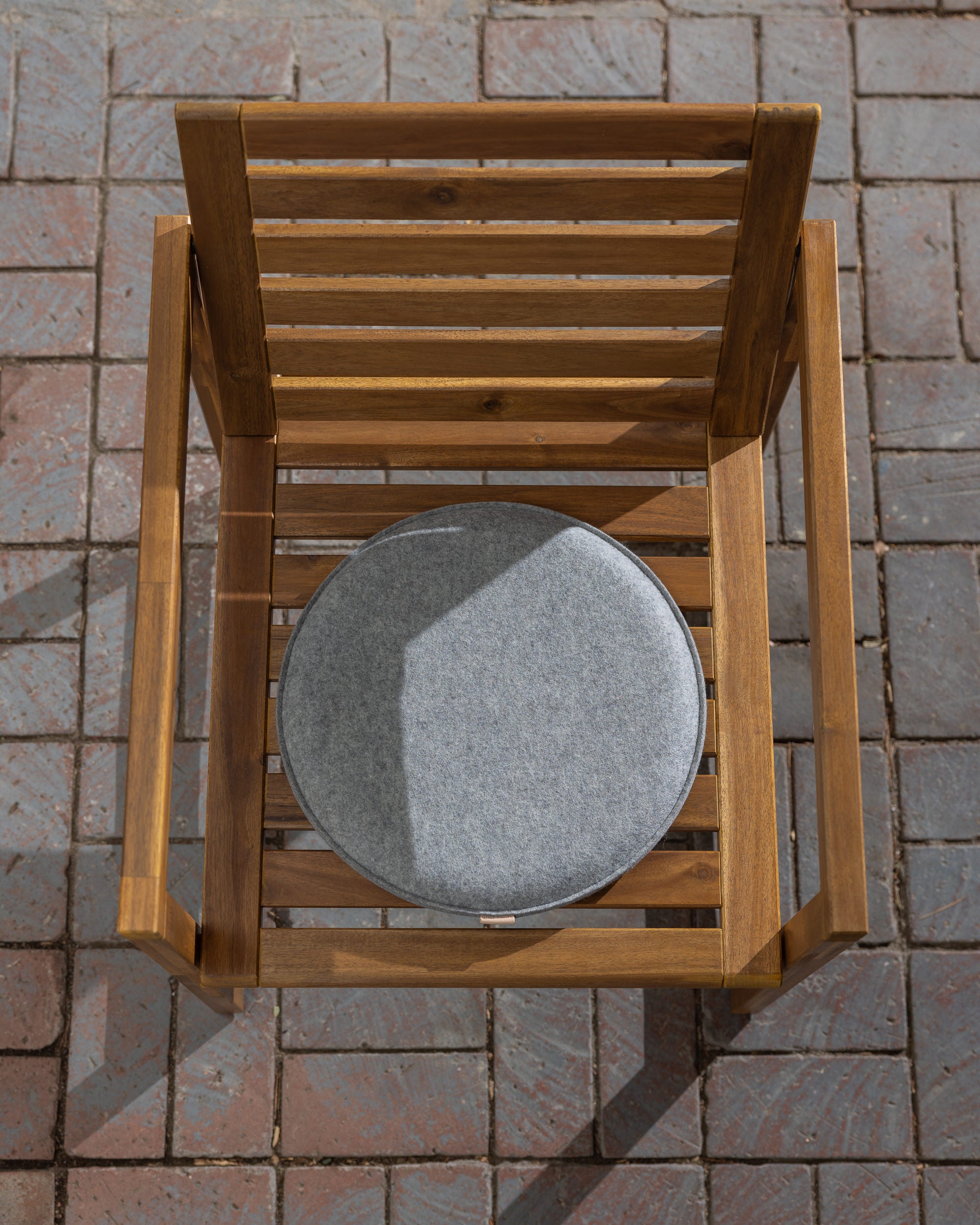 A top-down view shows a wooden chair with horizontal slats on a brick patio, topped with a round Merino wool cushion by graf lantz. Sunlight enhances the woods natural texture and casts a shadow, creating an ideal meditation spot.