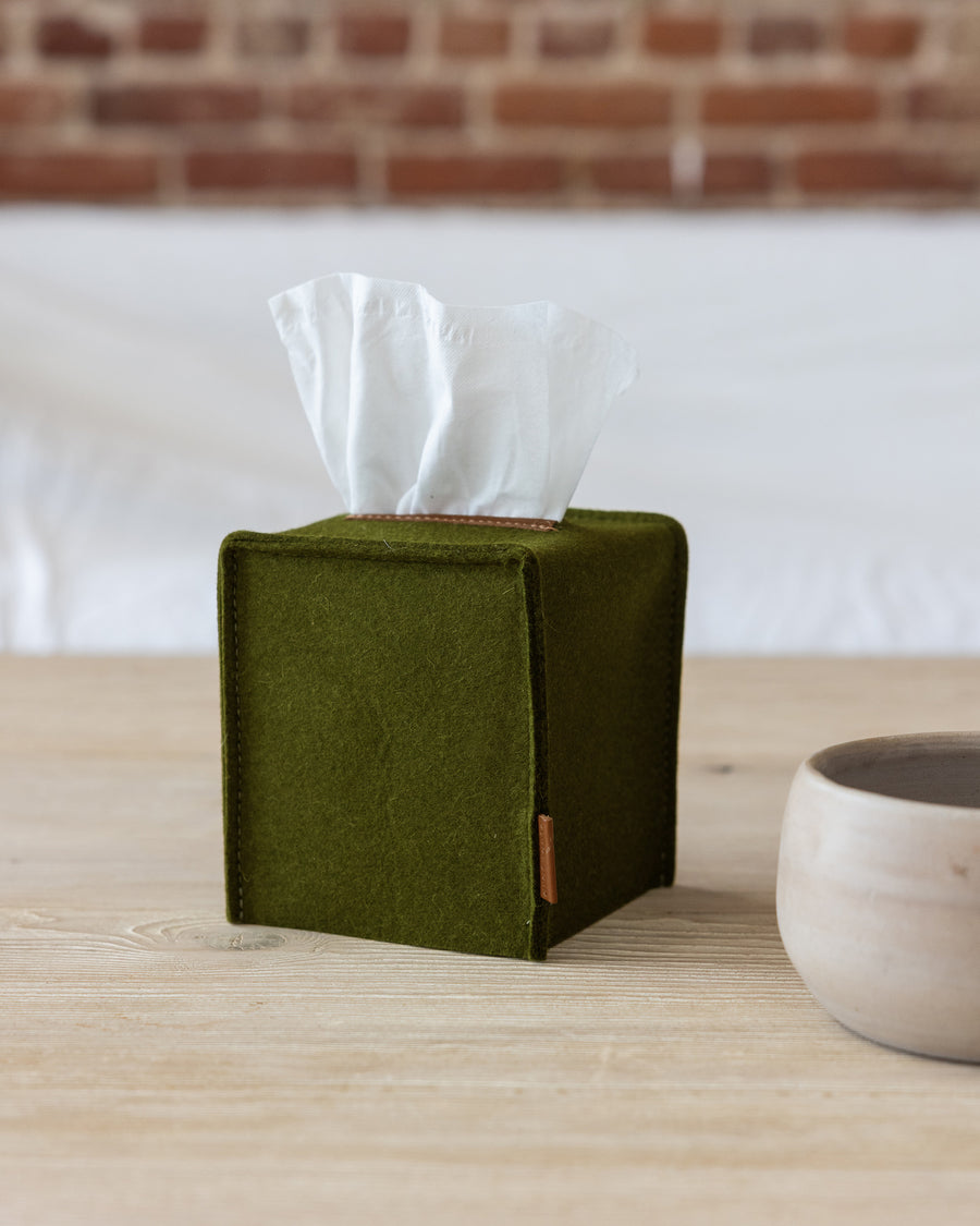 A small Merino Wool Felt Tissue Box Cover covering a square tissue box in color Moss Sienna with leather applications on a wooden tabletop next to a ceramic bowl