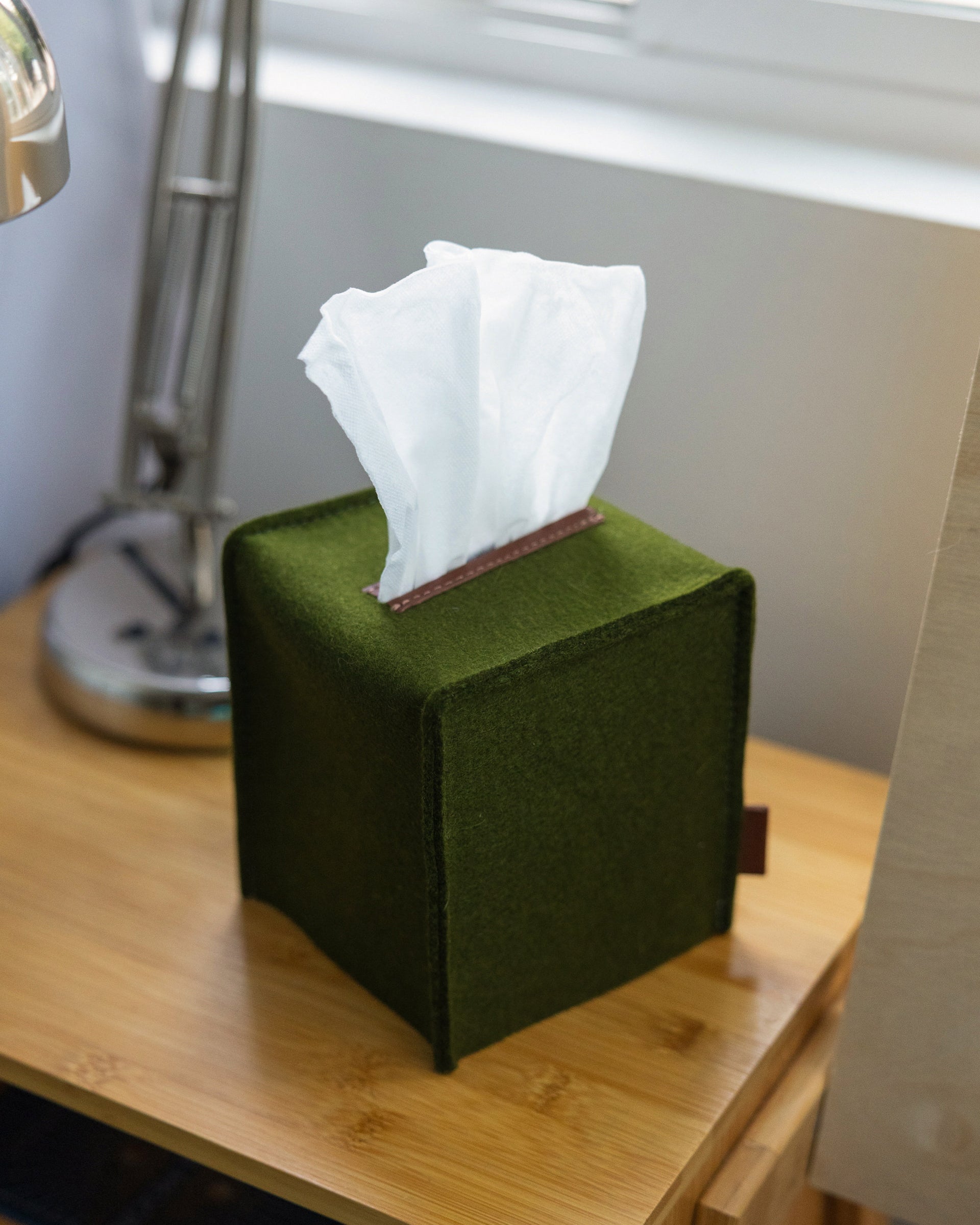 A Small Merino Wool Felt Tissue Box Cover covering a square tissue box in color Moss Sienna by Graf Lantz on a wooden nightstand next to a table lamp