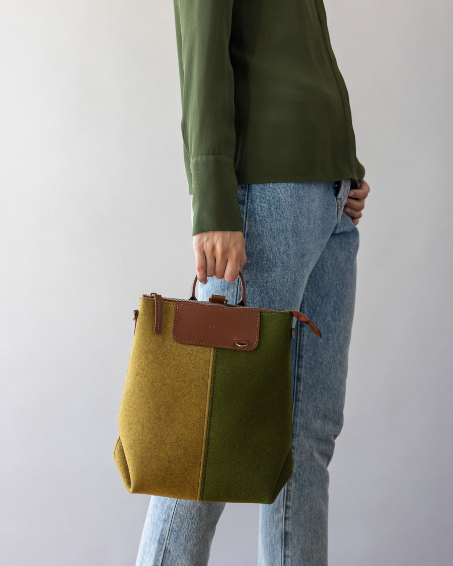 A woman holding a green and sand colored Bedford Merino Wool Colorblock Midi Backpack in one hand by her side