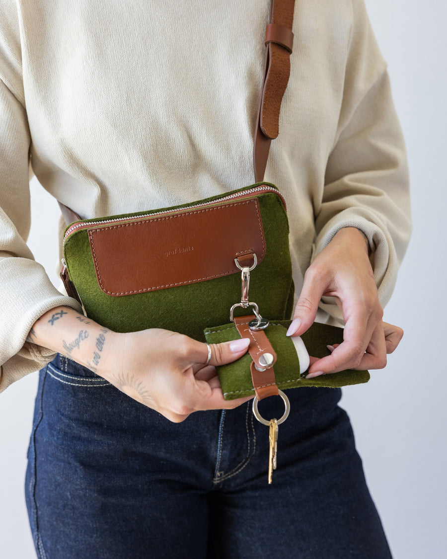 Standing woman with green Bedford Merino Wool Belt Bag used as crossbody with brown leather strap holding color-matching Merino Wool Pod Key Fob in both hands