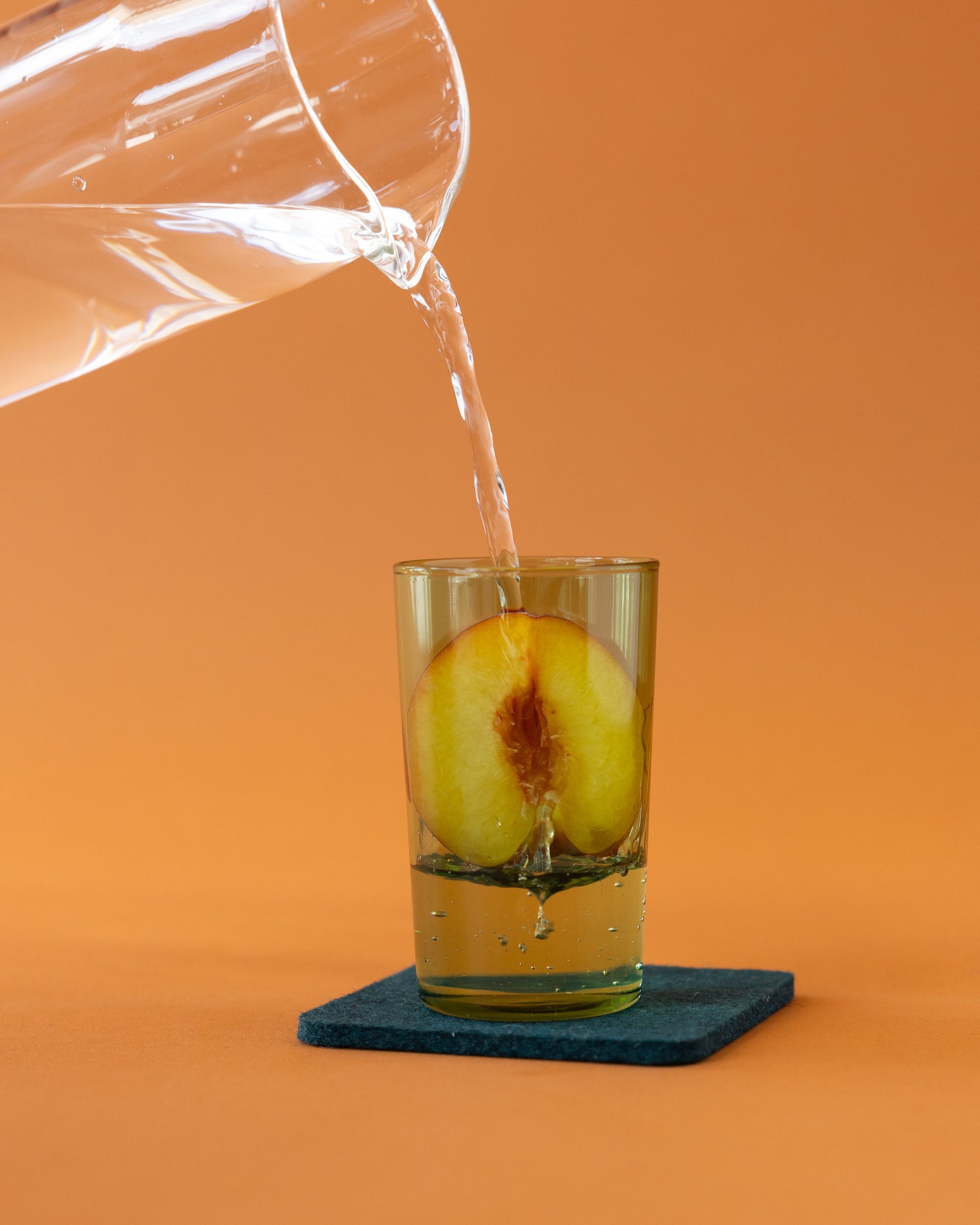 Clear pitcher pours water in one a cactus-colored glass with half peach inside, orange background