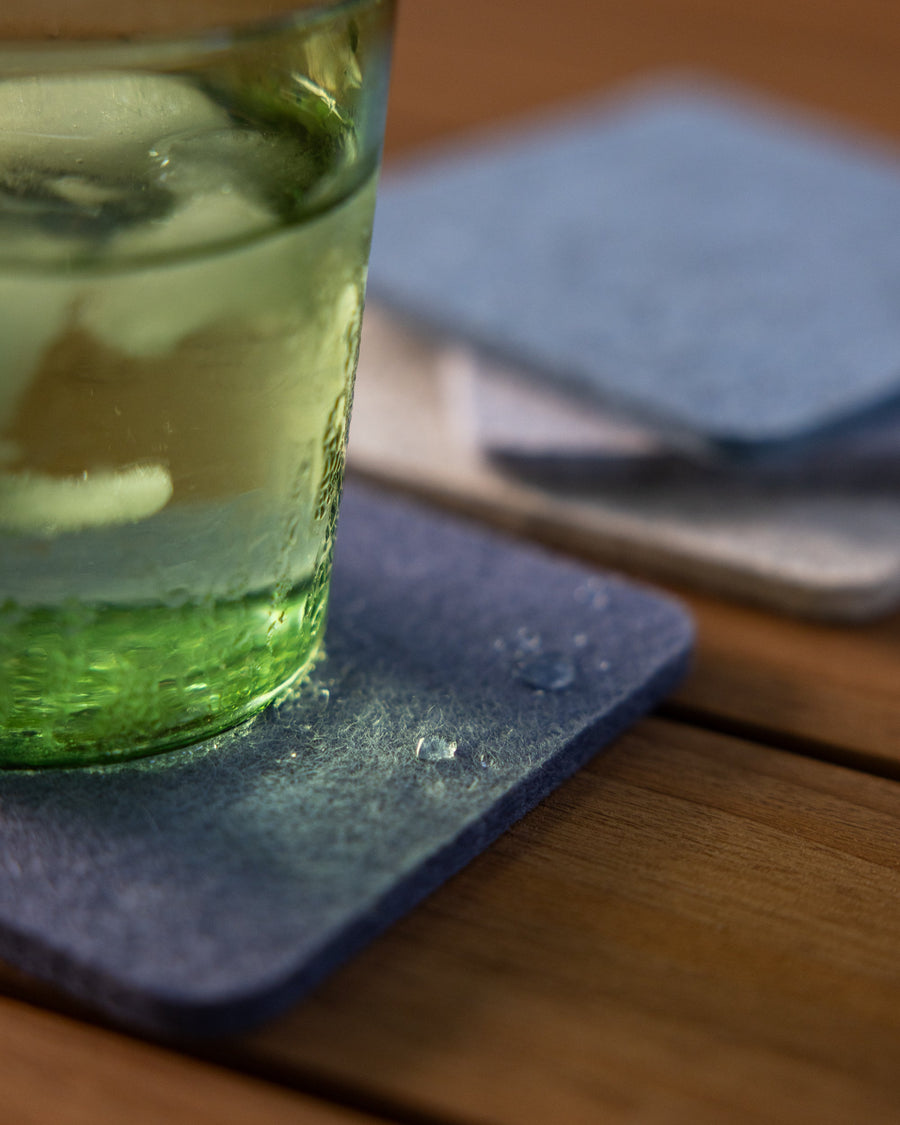 Close-up of a green drinking glass filled with water and ice cubes with condensation moisture on a square Merino wool coaster, three stacked coasters in background on wooden tabletop