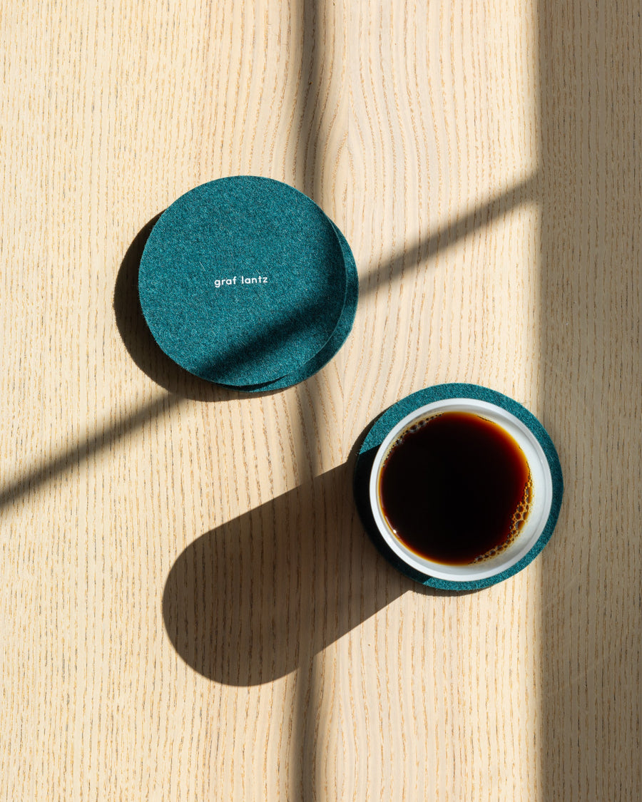 Four round coasters in color Kombu on a wooden tabletop, white drinking glass filled with coffee, view from above