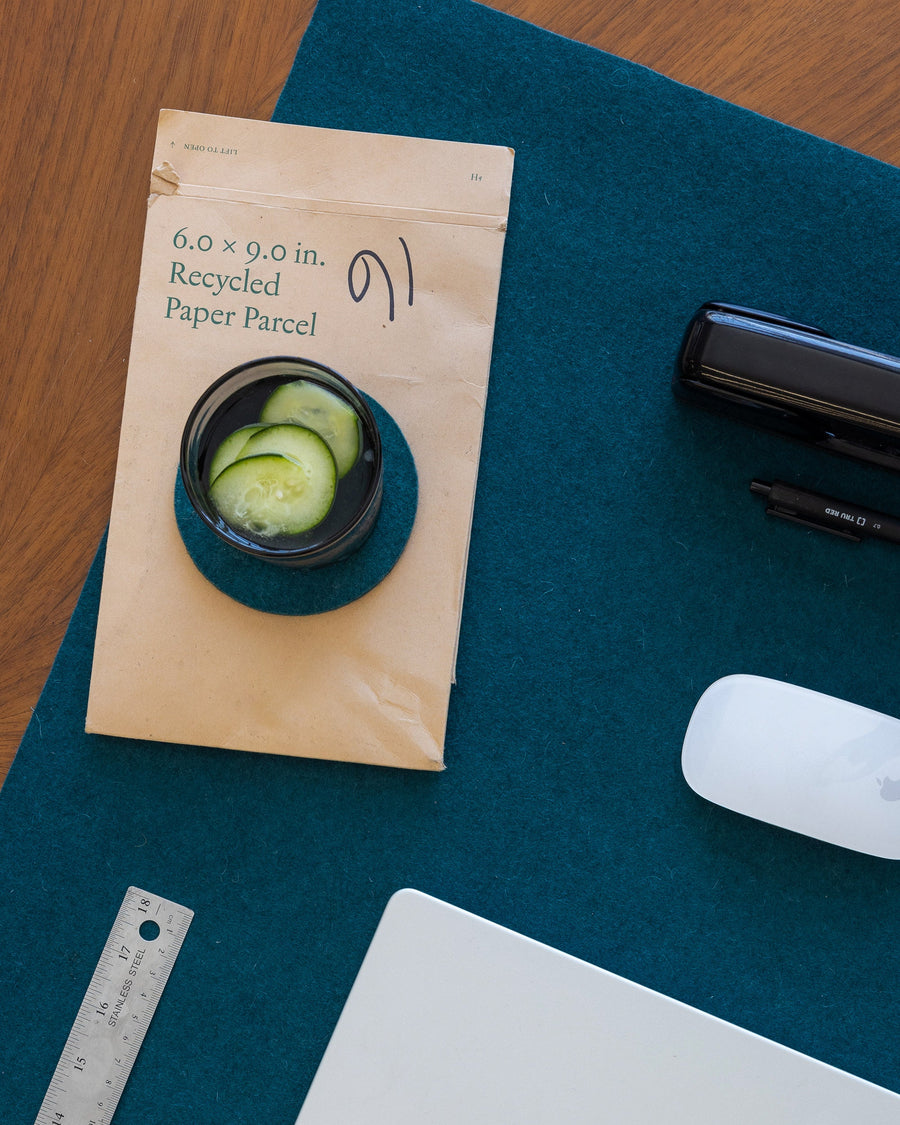 Glass on round Kombu-colored coaster, brown mailing envelope and kombu desk pad next to office accessories