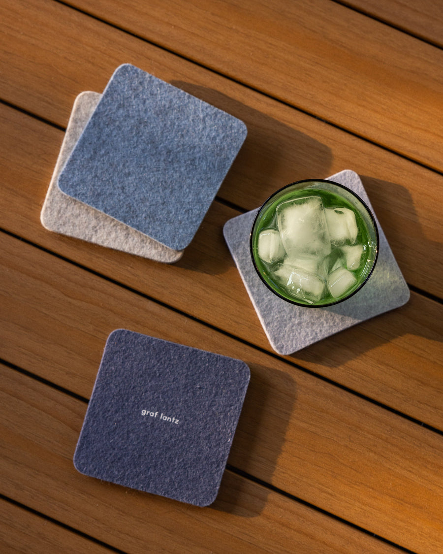 Set of Bierfilzl Merino Wool Square Coaster Multi 4 Pack in Heather Blue, Horizon, Fog, and Heather White as well as a green drinking glass on a wooden tabletop