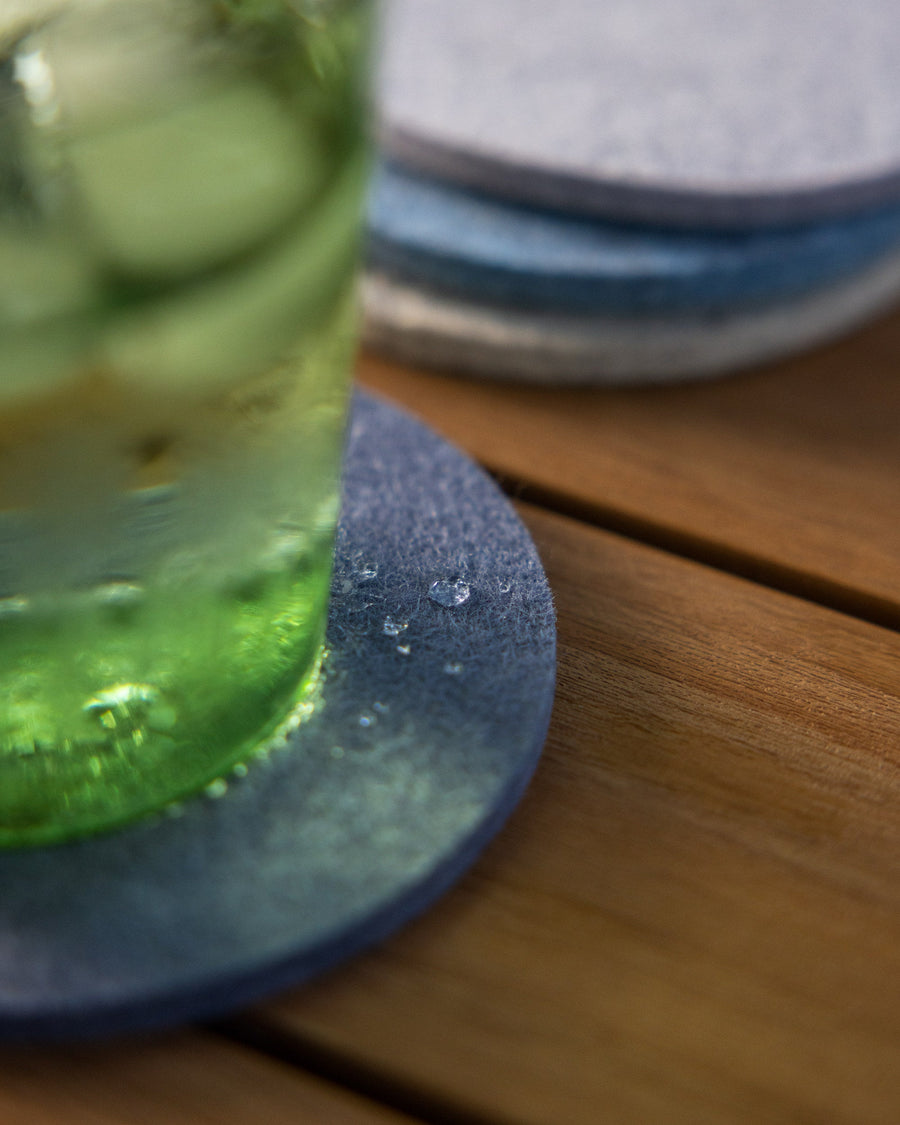 Close-up of a green drinking glass filled with water and ice cubes with condensation moisture on a round Merino wool coaster, three stacked coasters in background on wooden tabletop