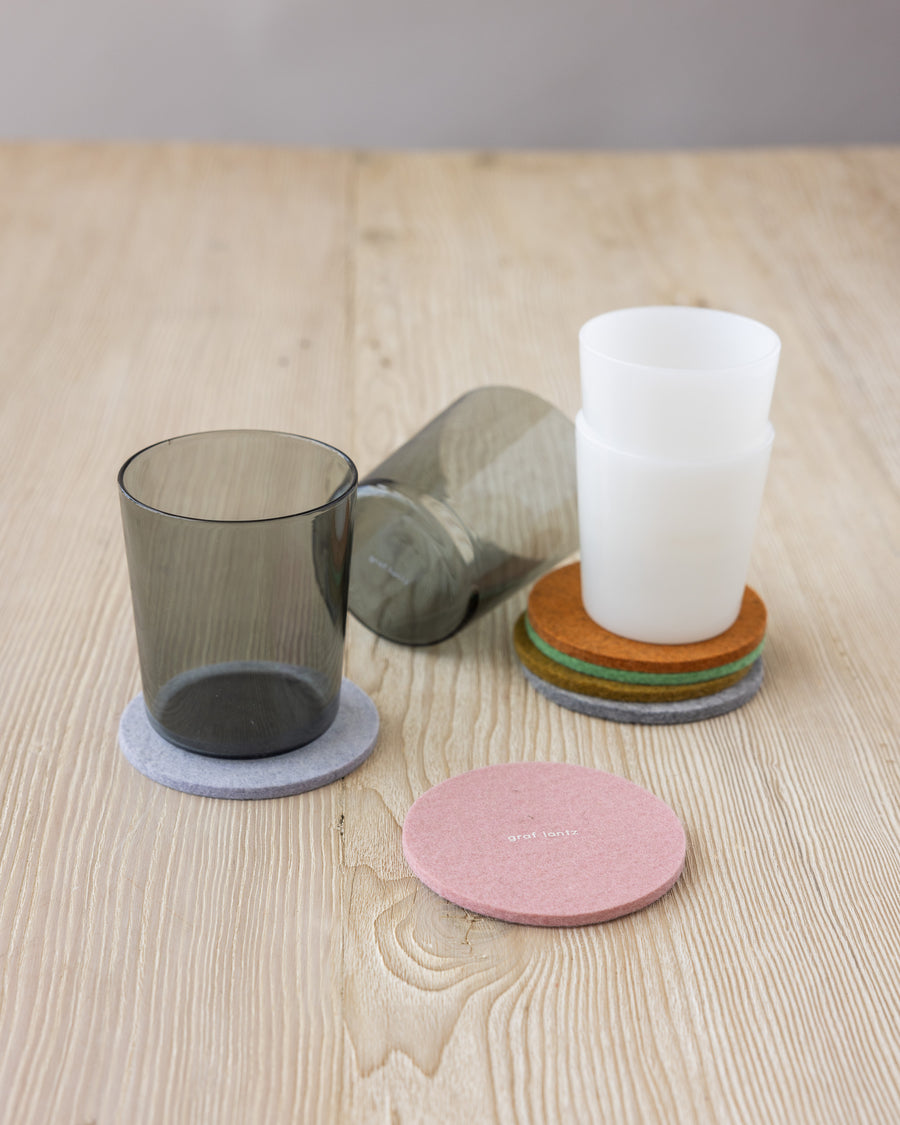 Stack of four round felt coasters by Graf Lantz in different colors on a wooden tabletop next to black and white glasses and two pastel-colored coasters