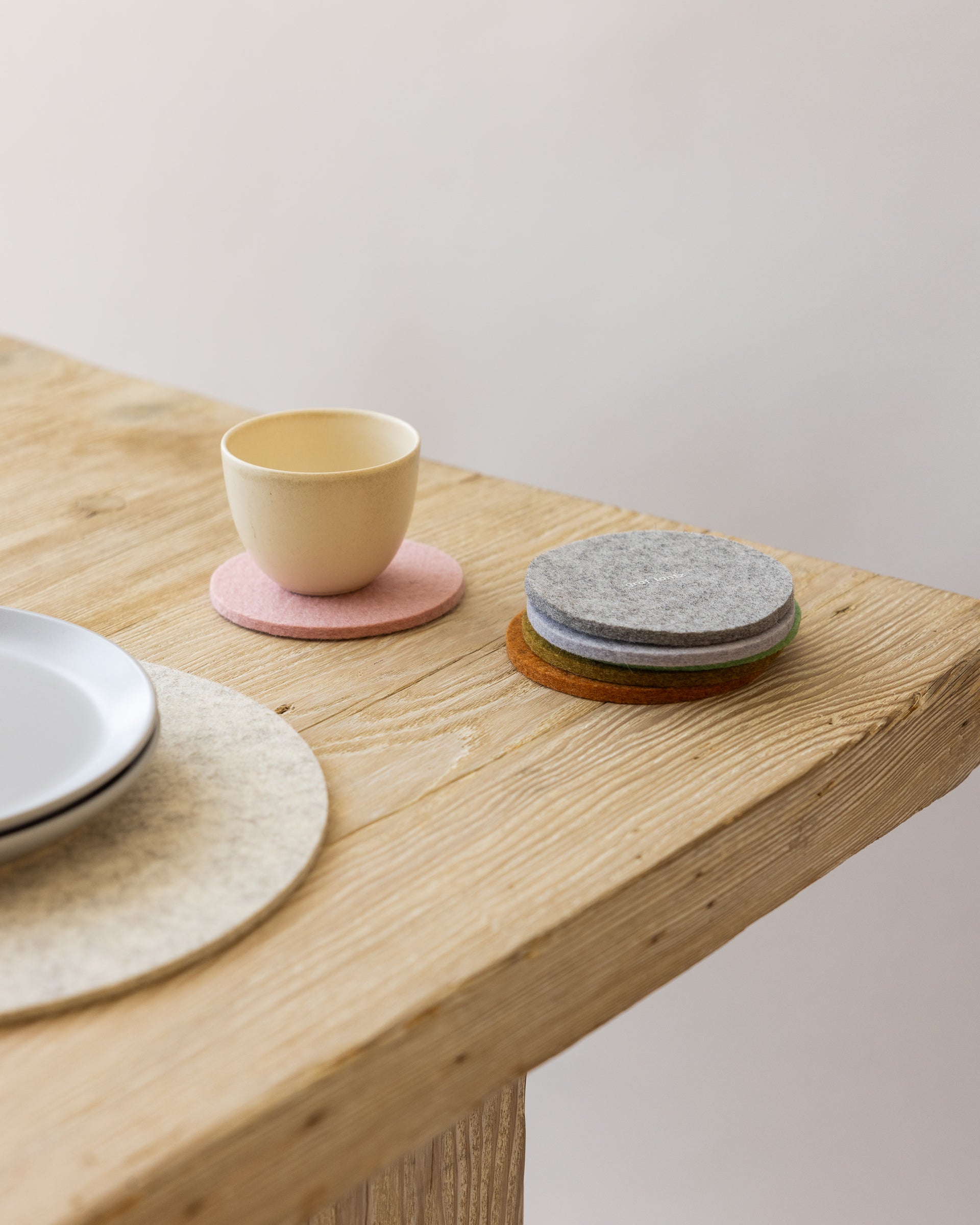 Stack of four round felt coasters in different colors on a wooden table next to a white placemat, white plates, and a small beige mug