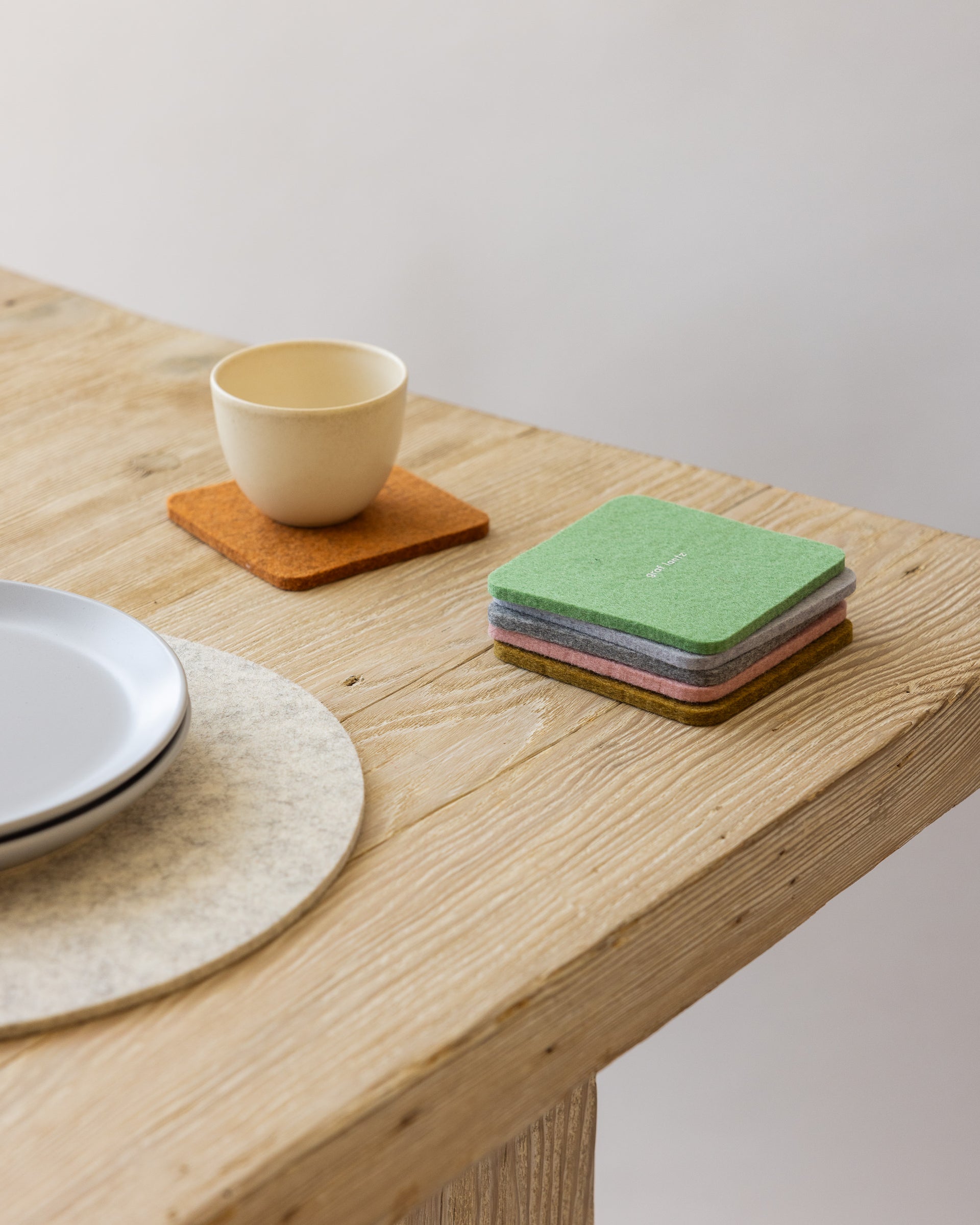 Stack of five felt coasters in different colors on a wooden table next to a white placemat, two stacked white plates, and a small beige mug on a coaster