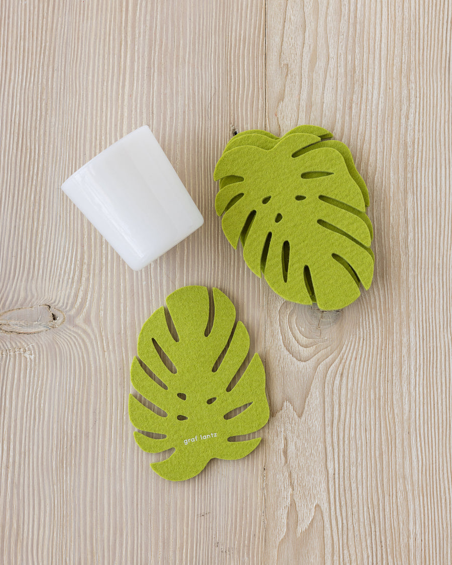 Two green Monstera welt coasters on a wooden tabletop next to a white glass