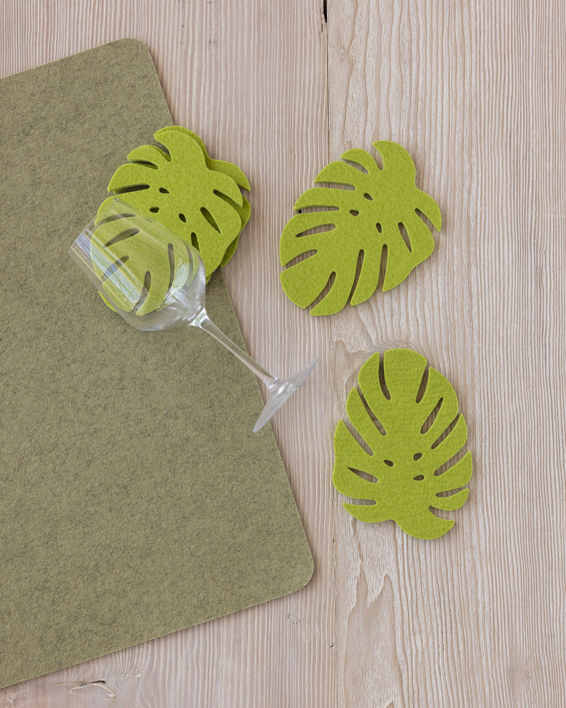 Three light green Monstera-shaped Merino wool coasters by Graf Lantz on a wooden tabletop on a green felt placemat next to a wine glass