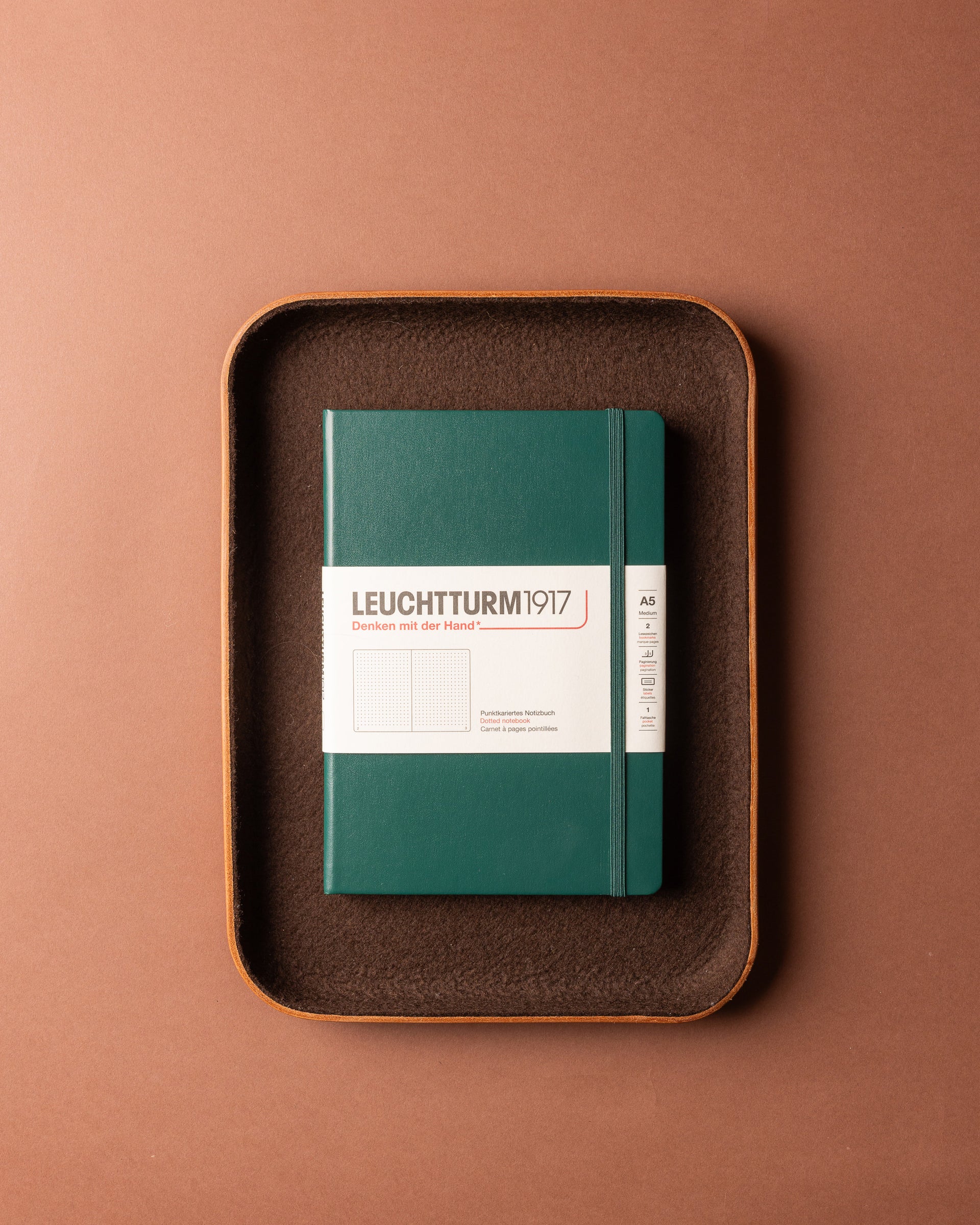 A green notebook with a white label sits centered on a brown Graf Lantz Merino wool tray, set against a solid brown background—ideal for organizing any workspace.