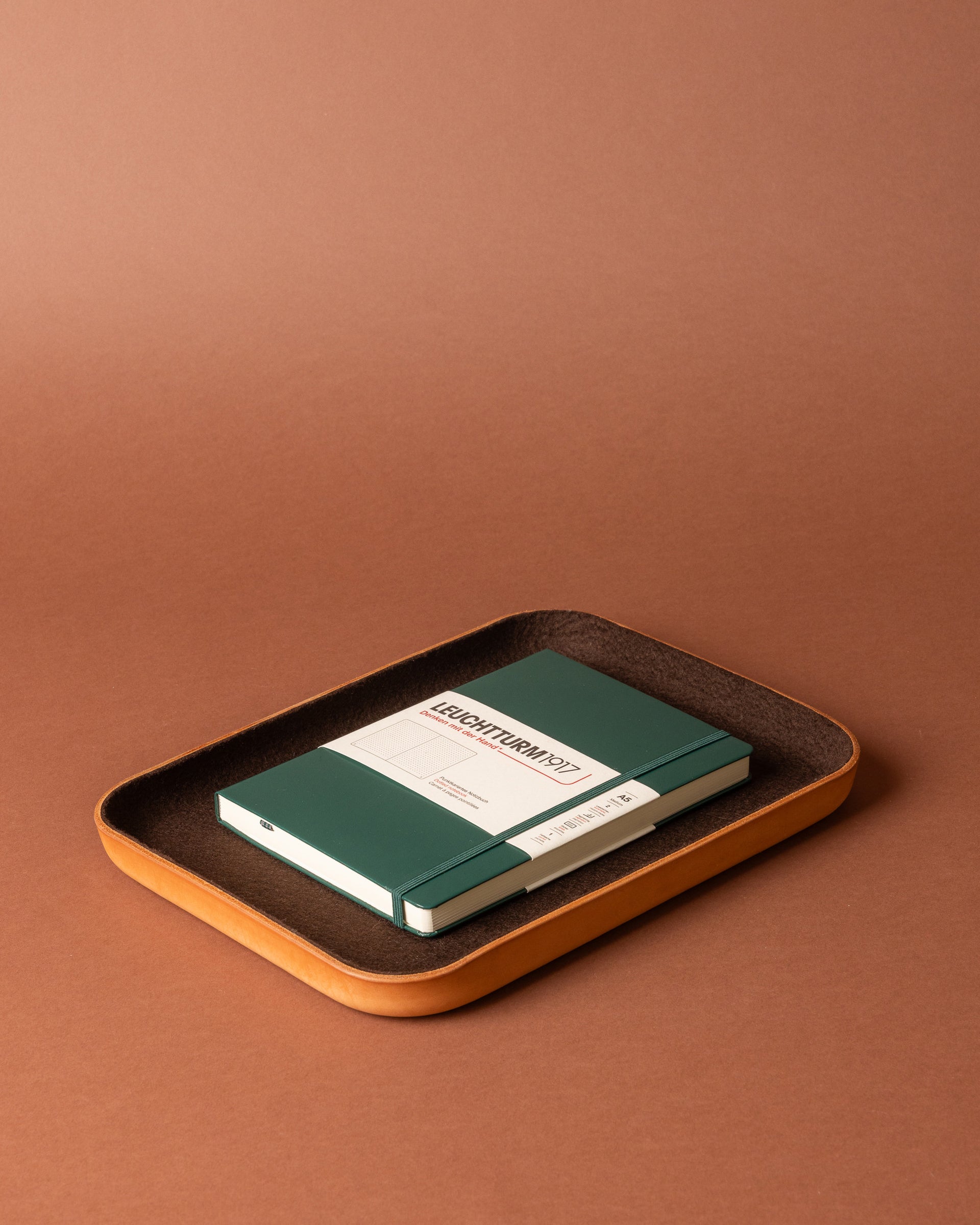 A green notebook and pen rest on a rectangular brown Merino wool tray from Graf Lantz, arranged on a matching brown surface and background for stylish workspace organization.