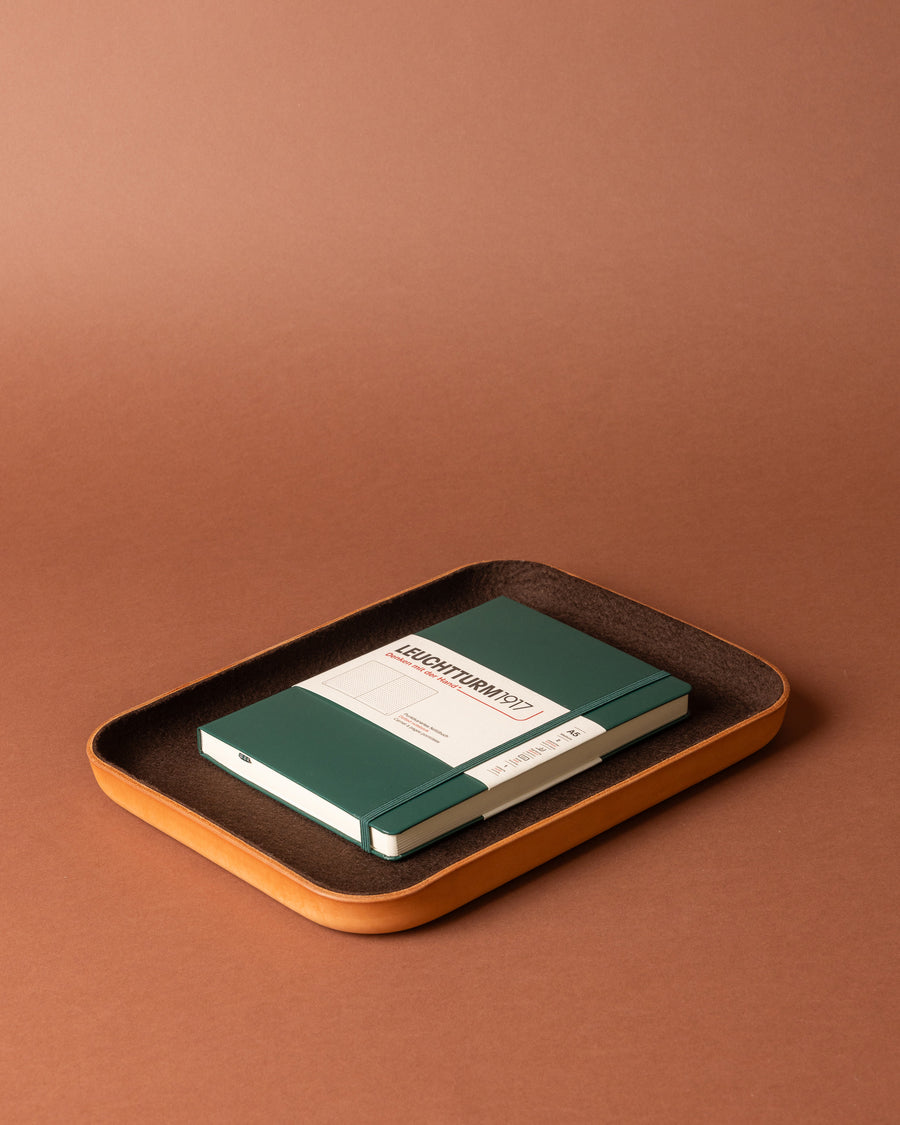 A green notebook and pen rest on a rectangular brown Merino wool tray from Graf Lantz, arranged on a matching brown surface and background for stylish workspace organization.
