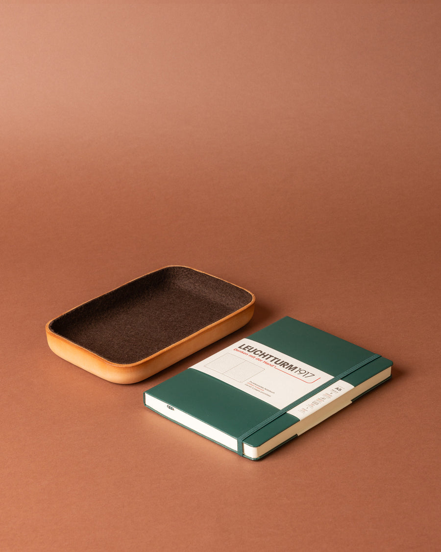 A rectangular brown Graf Lantz Merino Wool tray sits beside a closed green Leuchtturm1917 notebook with an elastic band, both arranged on a matching brown surface and background.