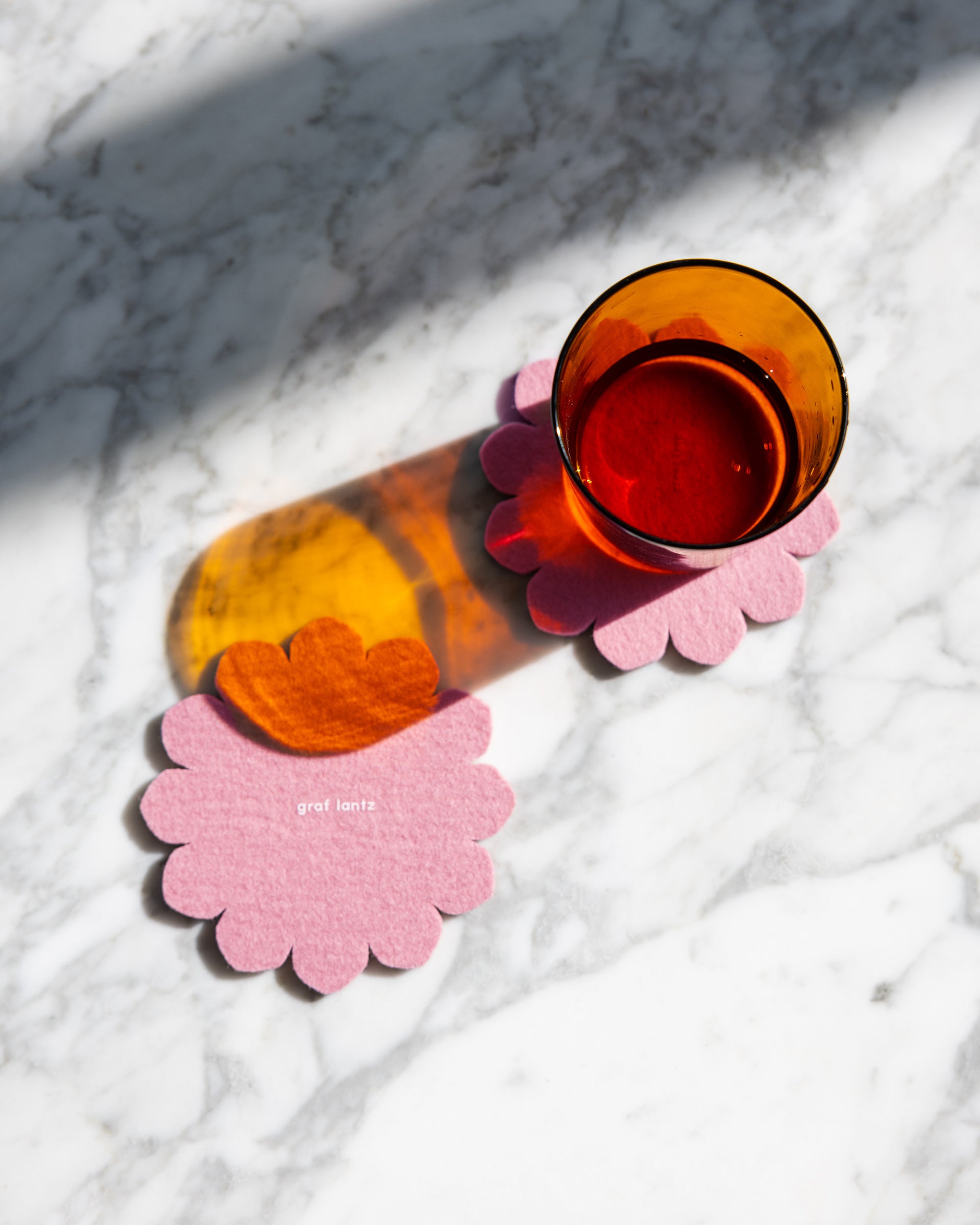 Two pink felt crest coasters by Graf Lantz on a marble tabletop, one orange-colored drinking glass standing on one of the coasters 