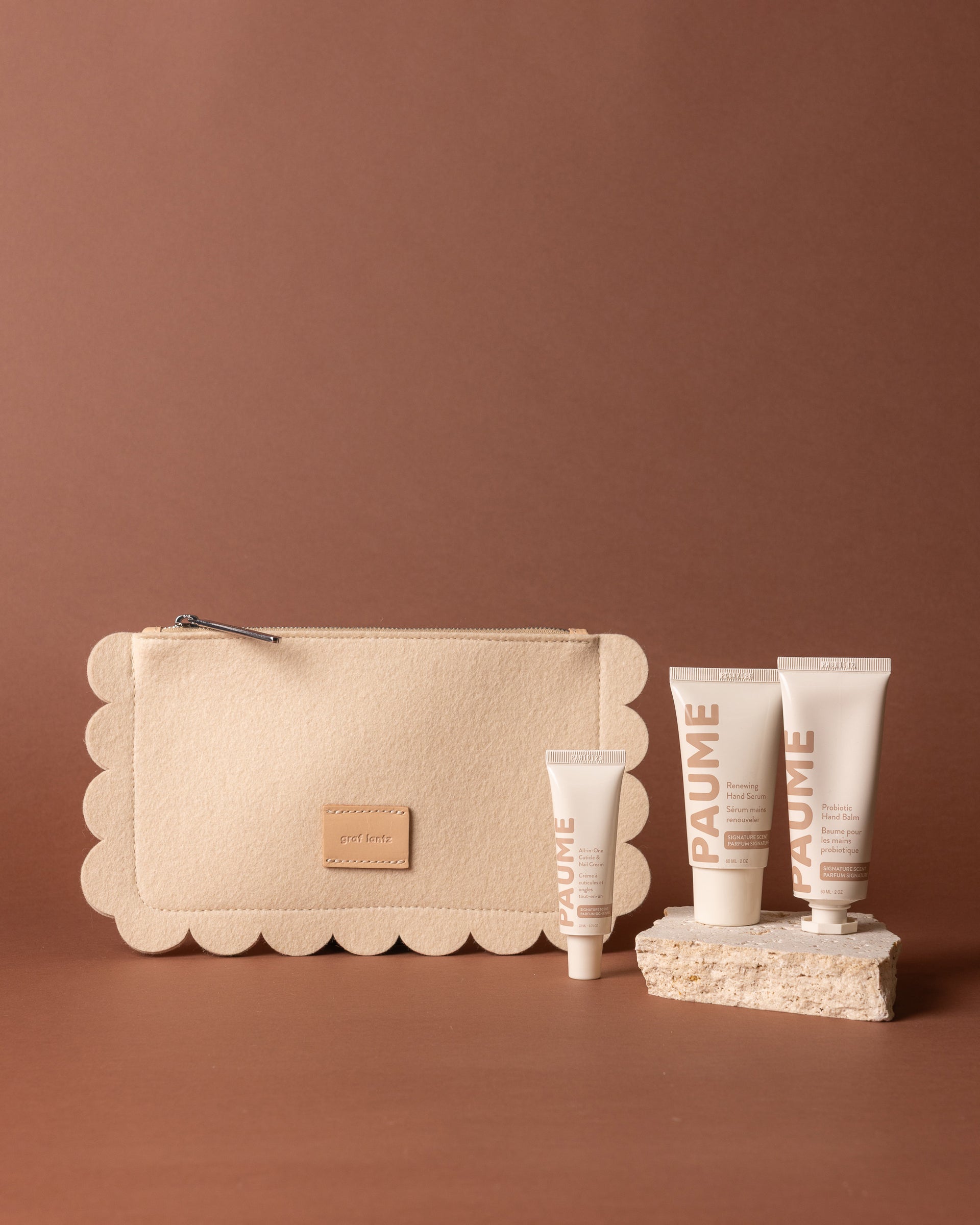 A beige Graf Lantz Merino wool pouch and four PAUME hand care tubes are displayed on a brown background, with three tubes upright on a textured stone and the smallest tube positioned in front.