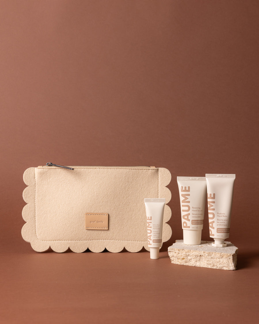 A beige Graf Lantz Merino wool pouch and four PAUME hand care tubes are displayed on a brown background, with three tubes upright on a textured stone and the smallest tube positioned in front.