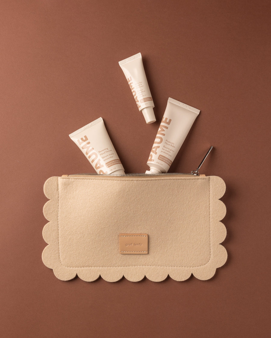 A beige Graf Lantz Merino wool pouch with scalloped edges lies on a brown background. Four Paume hand care tubes, including probiotic hand balm, are partially tucked inside, with three tubes sticking out at various angles.