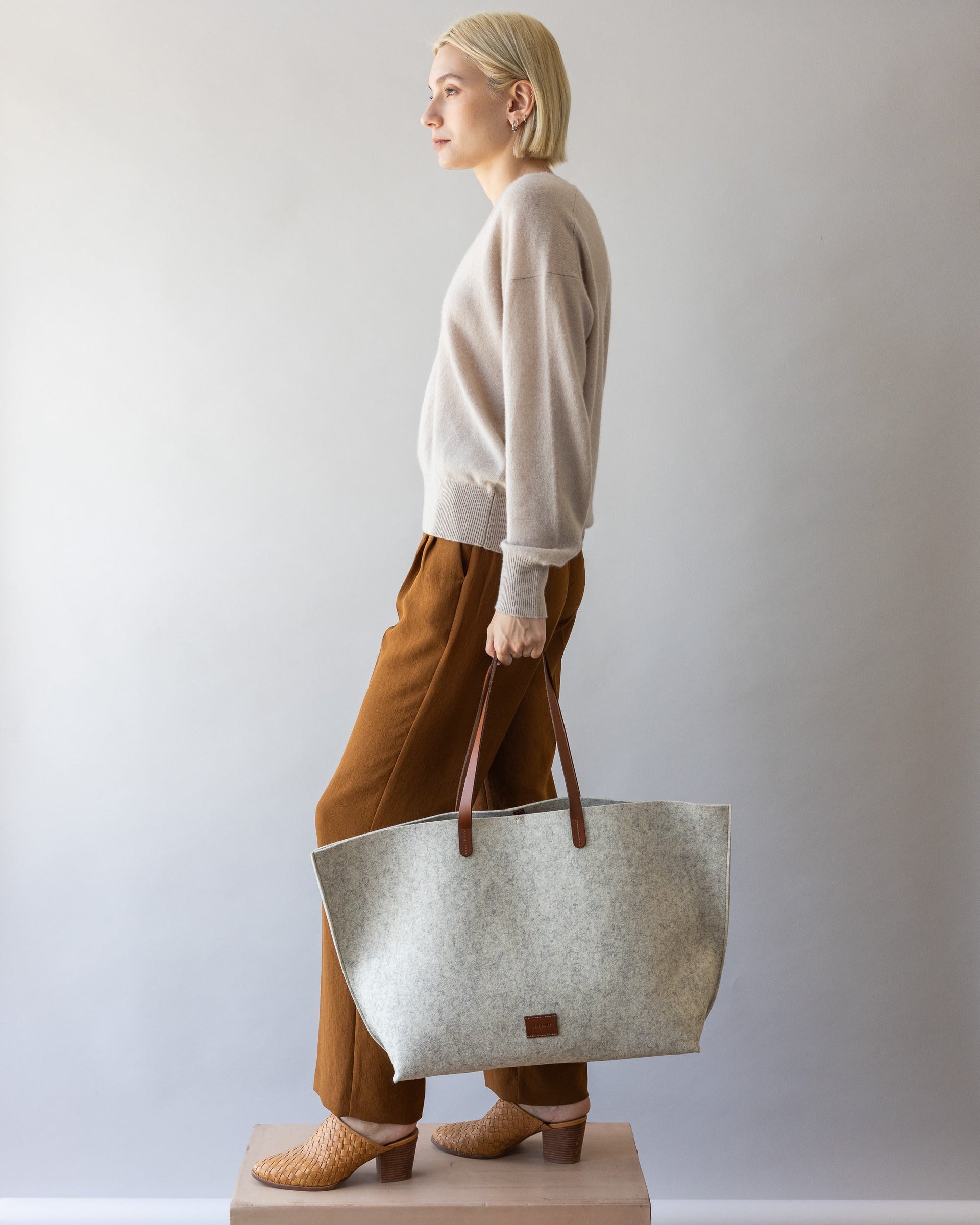 Standing woman holds brown leather straps of White Hana Merino Wool Boat Bag in one hand by her side, side view.