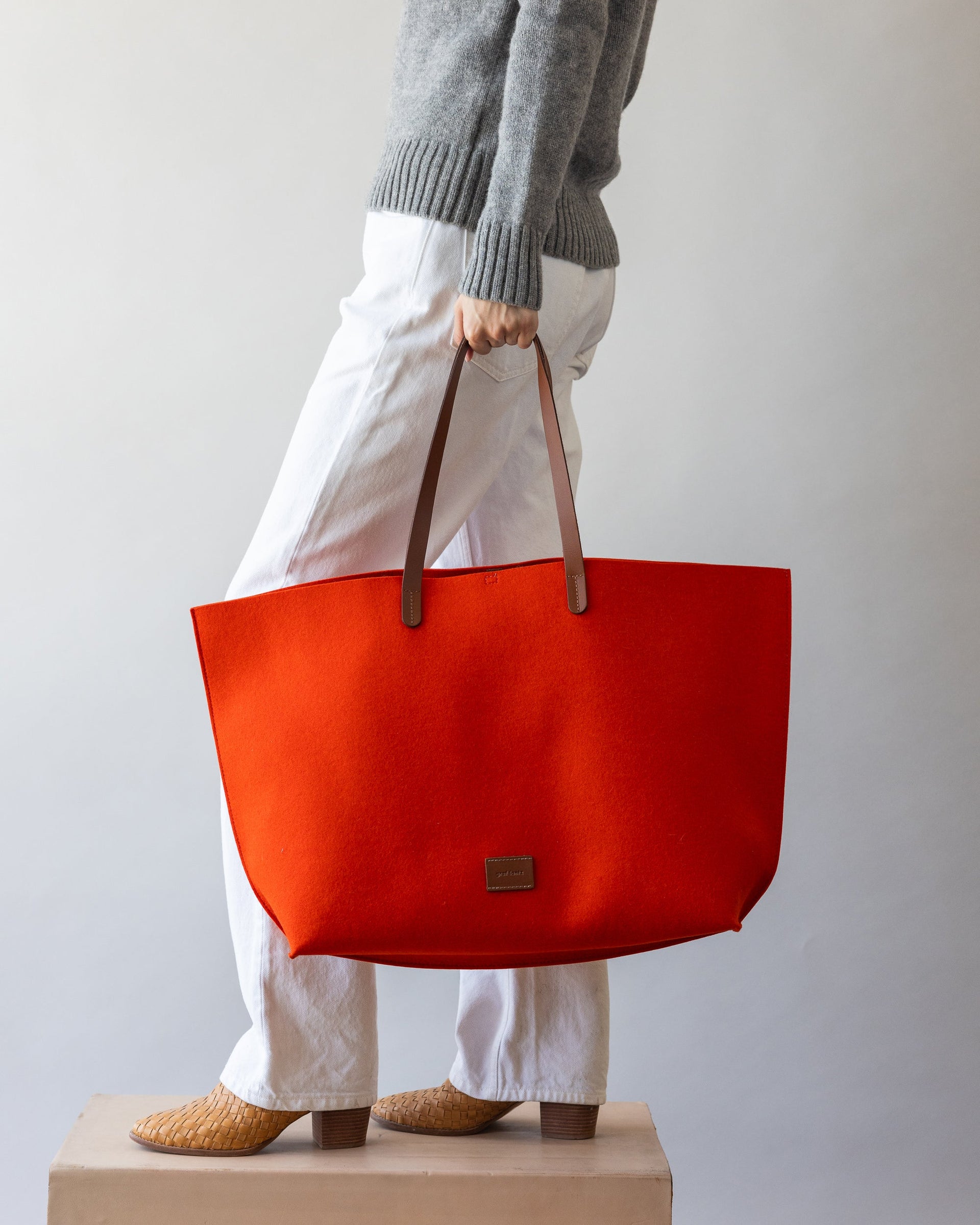 Standing woman holds brown leather straps of orange Hana Merino Wool Boat Bag in one hand by her side, side view.