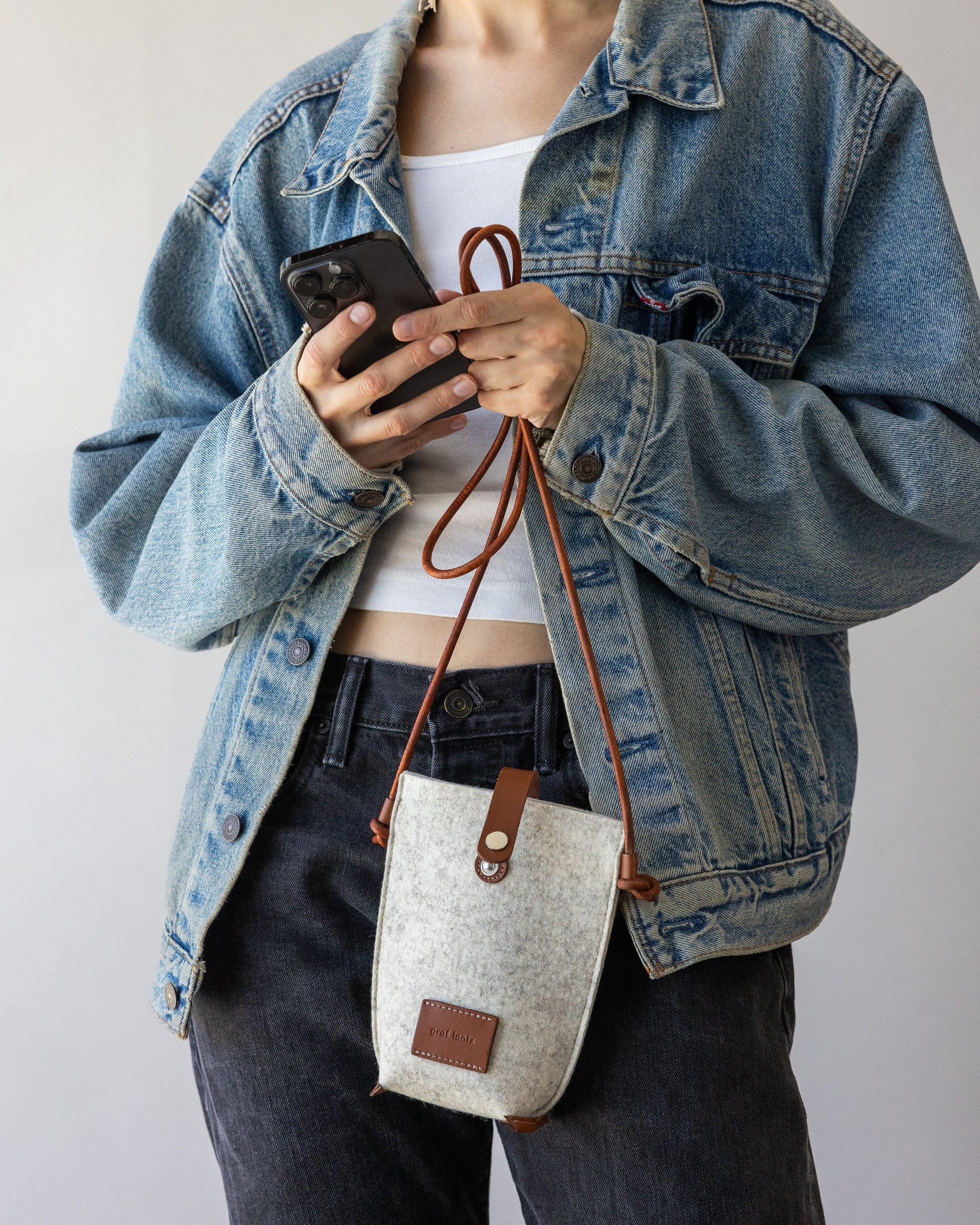 Woman holds brown leather strap of Hana Merino Wool Phone Crossbody in heather white in one hand, a black mobile phone in the other hand.