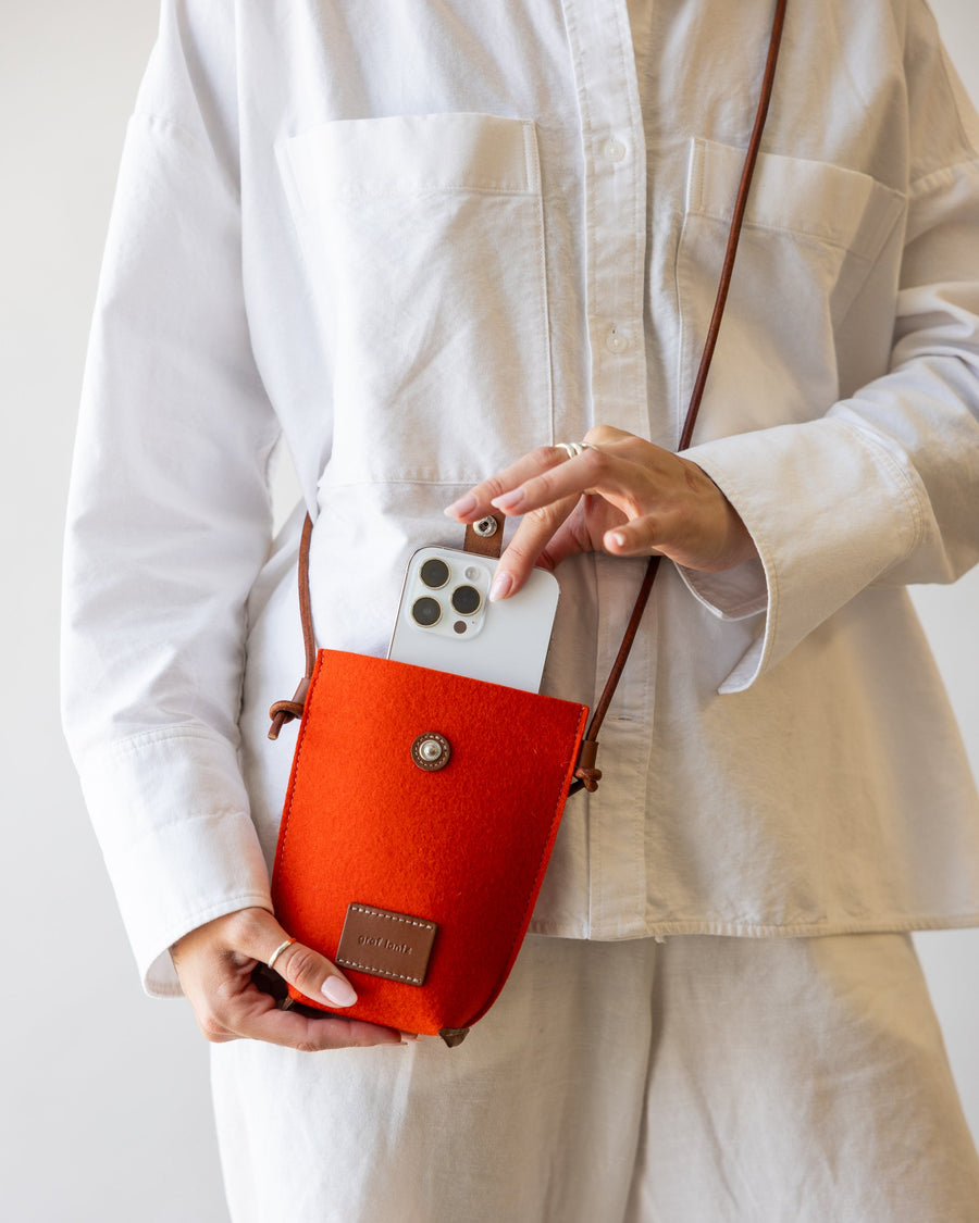 Woman holds orange Hana Merino Wool Phone Crossbody in one hand while the other hand opens the snap closure to grab the mobile phone inside.