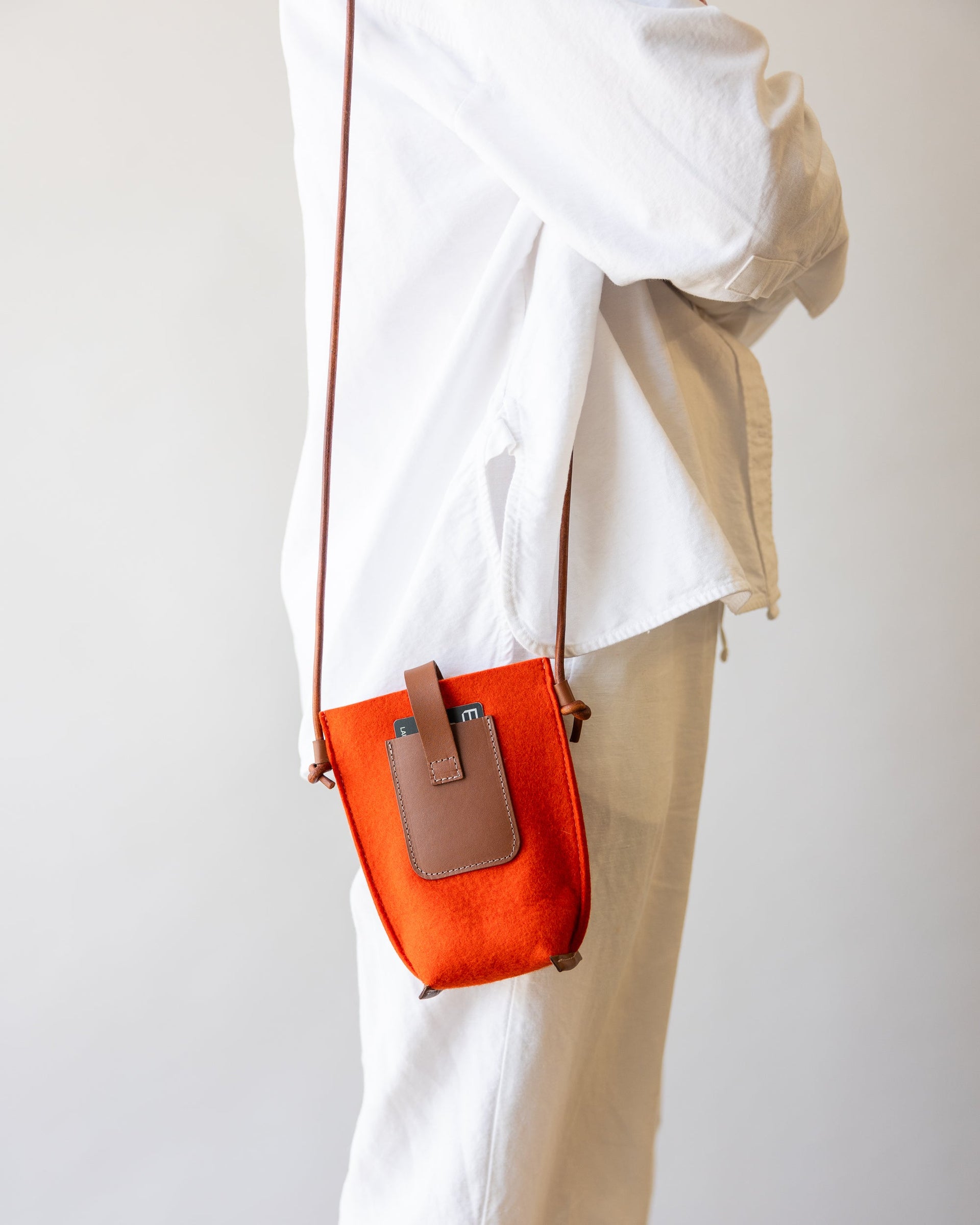 Standing woman dressed in white with dark brown straps of orange Hana Merino Wool Phone Crossbody bag over one shoulder, side view.