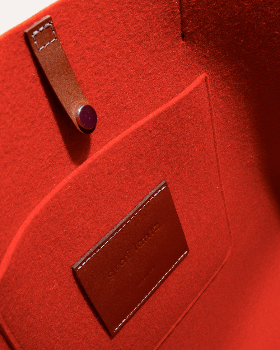 View in an orange Merino Wool Tote bag featuring a pocket, brown leather applications, and snap closure