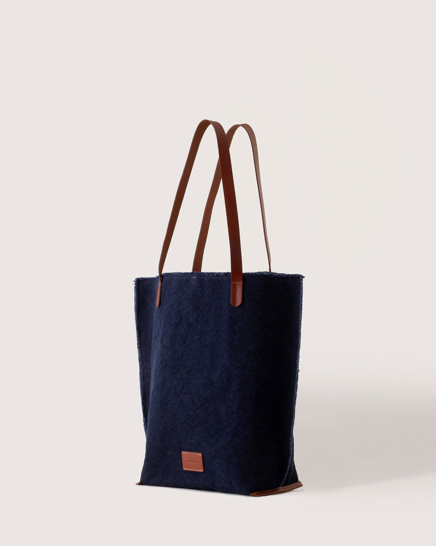 A dark blue Hana Canvas Tote with dark brown leather handle, white background, side view