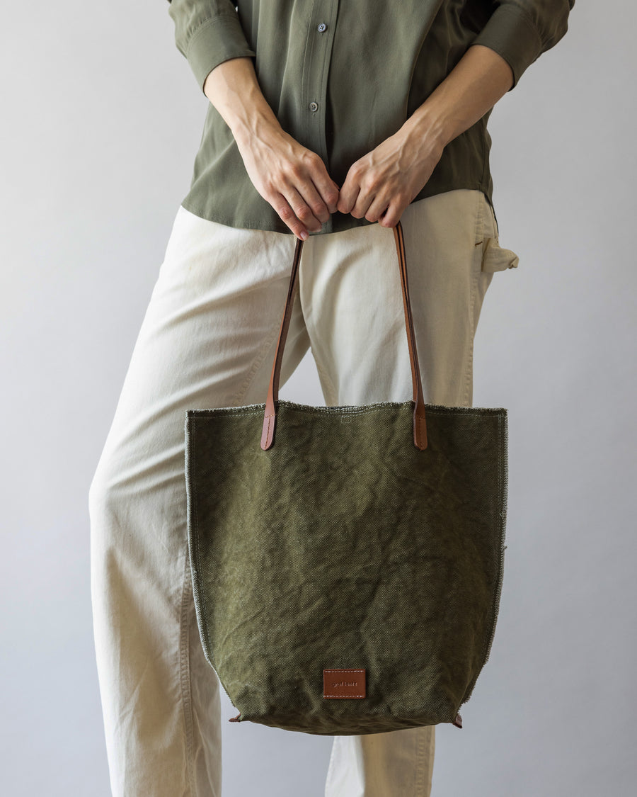 A woman holding a Hana Canvas Tote by Graf Lantz in Olive Sienna in front of her on the dark brown leather handle