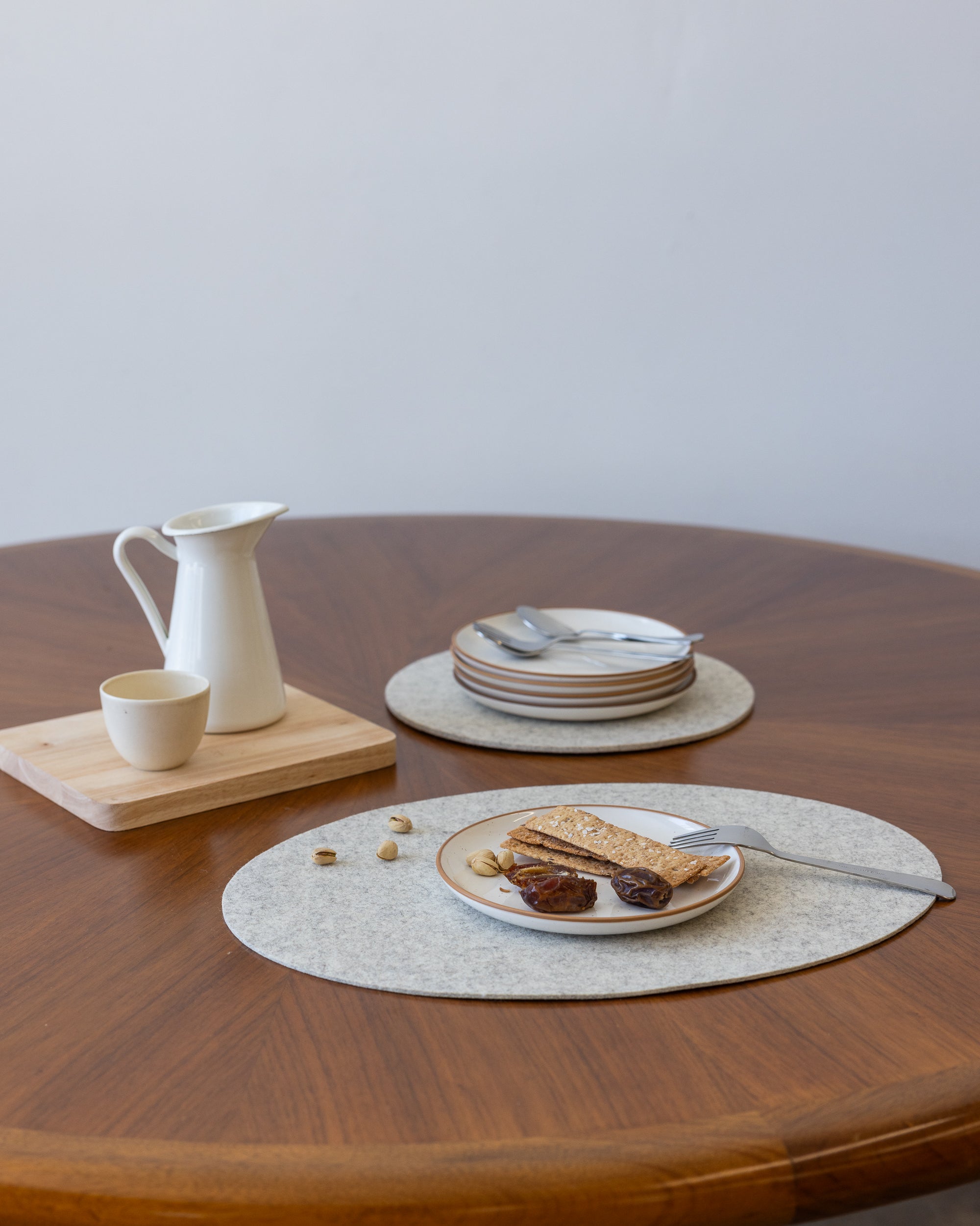A round table features a white pitcher and cup on a wooden tray, with a nearby stack of plates and cutlery ready for use. Toast, figs, and nuts are artfully placed on a heat-resistant Merino Wool Placemat by graf lantz in the foreground.
