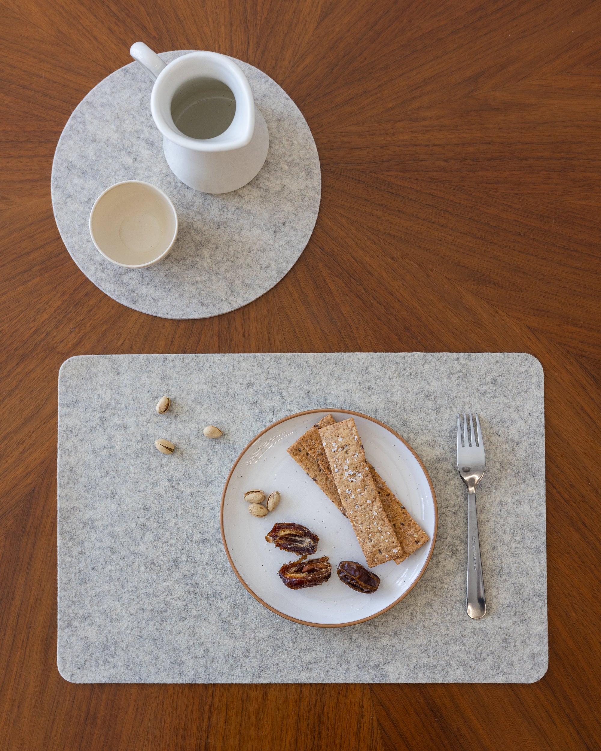 A wooden table showcases a graf lantz Merino Wool Placemat, famous for its moisture-wicking properties, holding a plate of crackers, dates, and pistachios. A fork rests next to it. Above, a heat-resistant coaster supports a white pitcher and an empty cup.