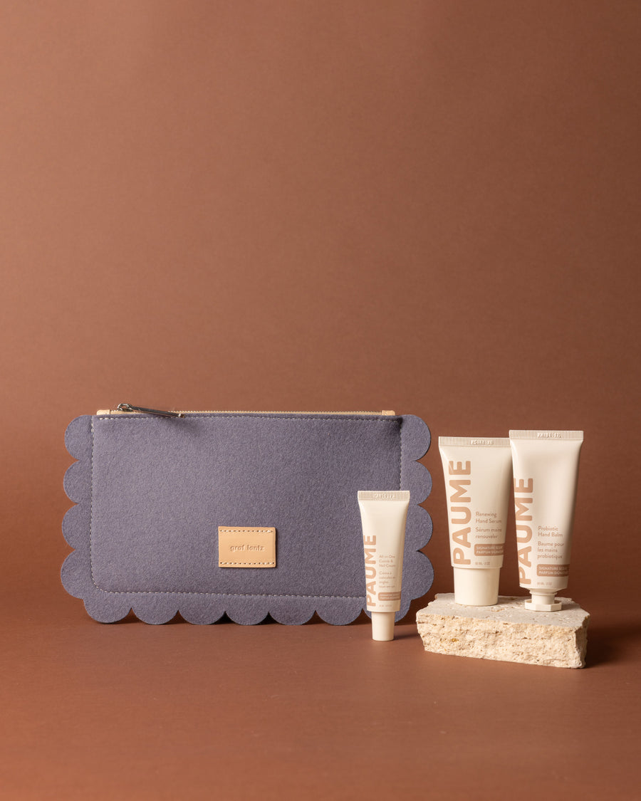 A Horizon blue, scalloped-edge Graf Lantz Merino Wool Pouch sits next to four beige PAUME Hand Care product tubes on a brown background, with some Probiotic Hand Balm tubes elevated on a light stone.