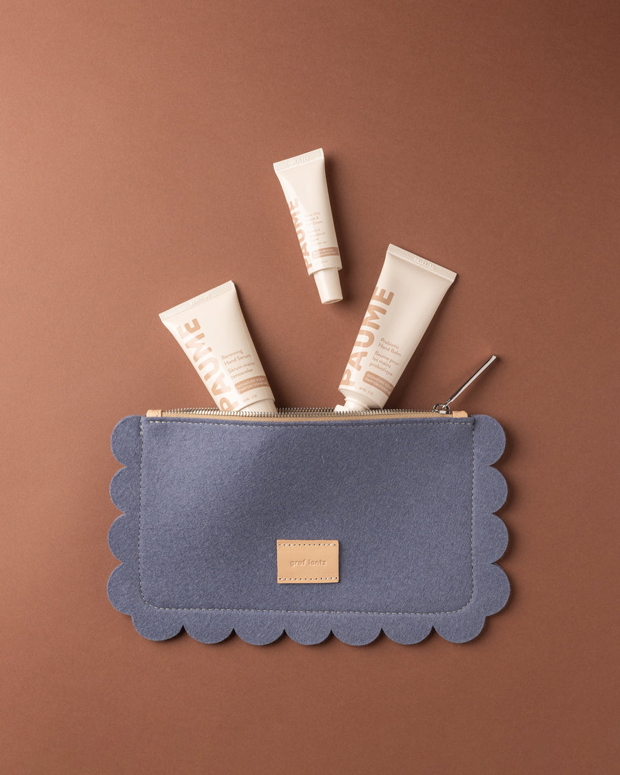 A Horizon blue scalloped-edge Merino Wool Pouch by Graf Lantz with a zipper is partially open, revealing three cream-colored tubes of Paume Hand Care products against a brown background.