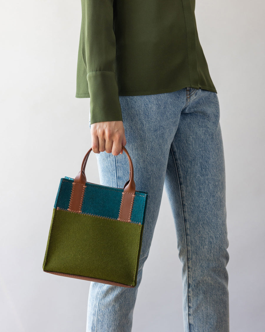 Standing woman holds Moss and Kombu colored Jaunt Merino Wool Mini Tote in one hand by her side, white background