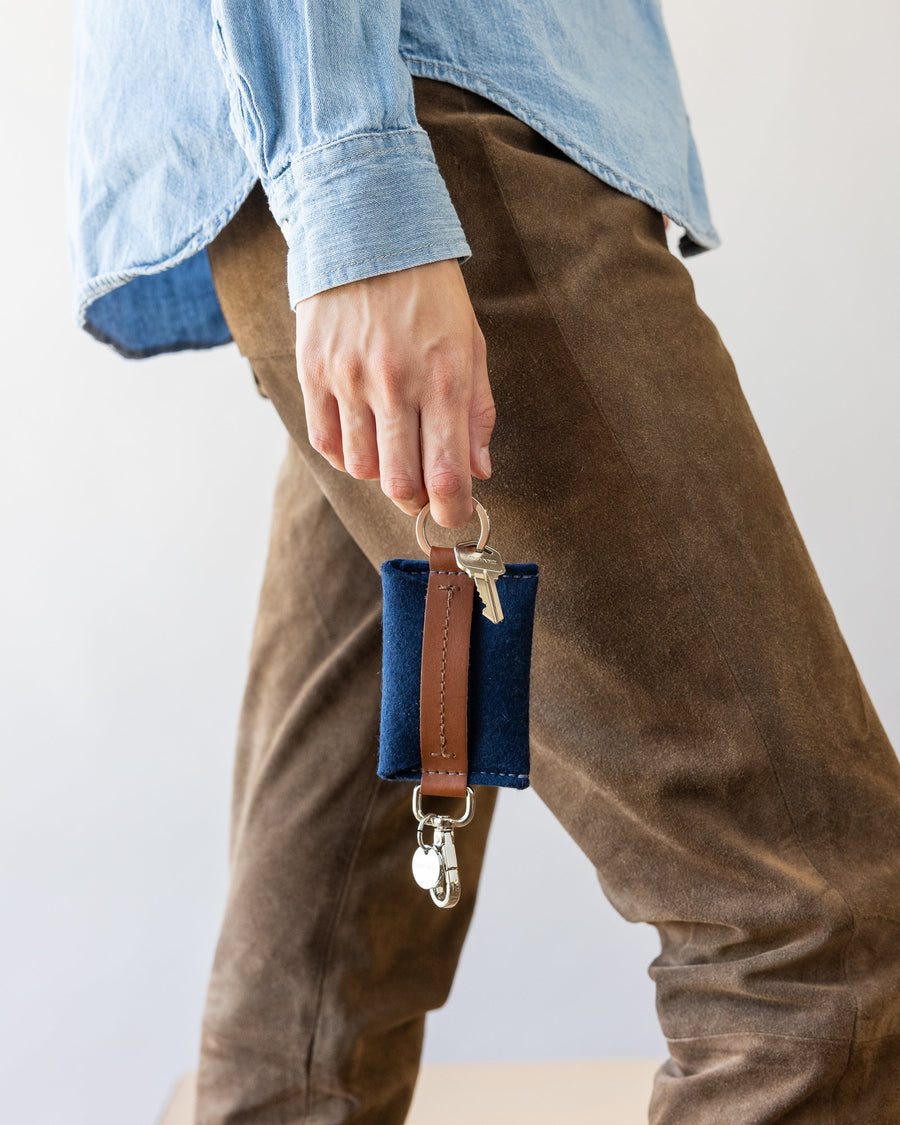 Walking woman holds Marine-colored Merino Wool Card Key Fob on one finger beside her leg in brown pants, white background.