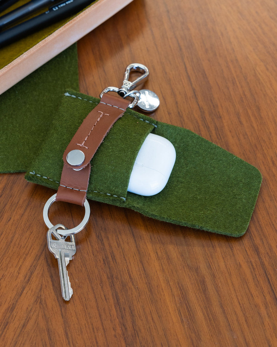 An open green Merino Wool Pod Key Fob with brown leather accents with a white pod case on a wooden tabletop