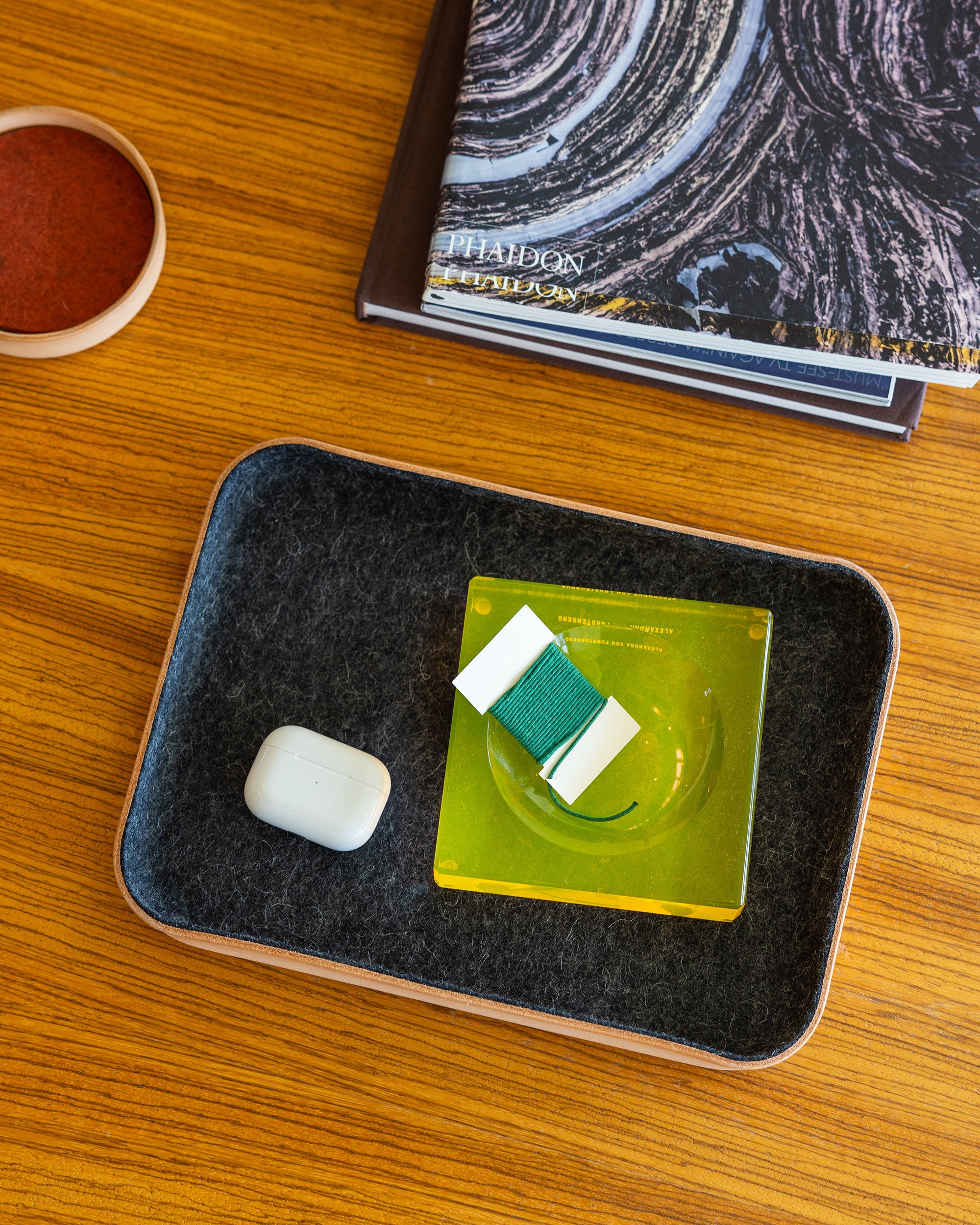 A wooden table showcases a Vachetta leather tray by graf lantz, holding a white earbud case and a green square item with small white strips. Nearby lies a closed book with a swirling cover pattern and a small round container.